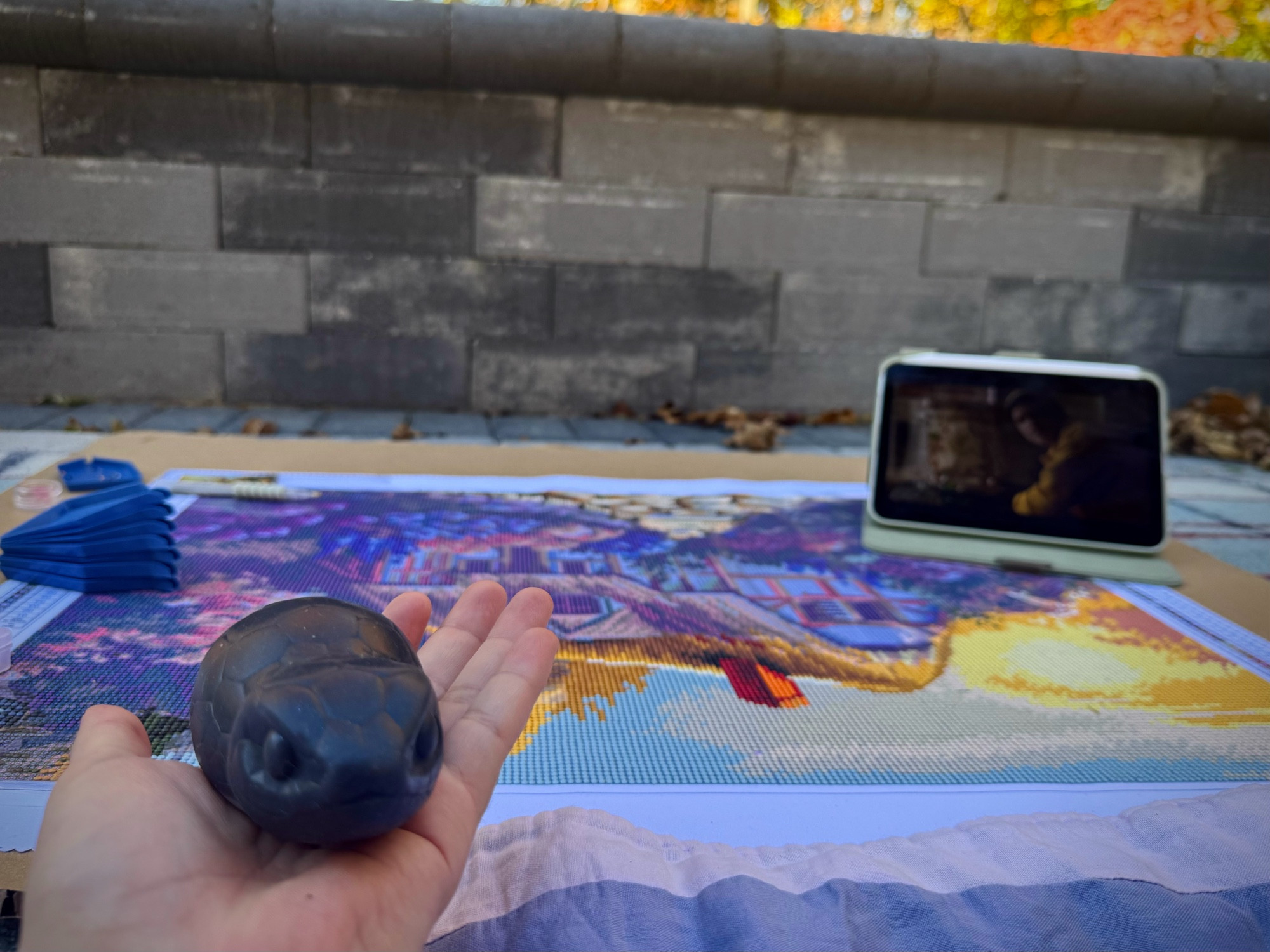 A photo of my hand, holding a squishy tsuchinoko, sitting outside with a diamond art kit in front of me while Netflix plays in the background. 