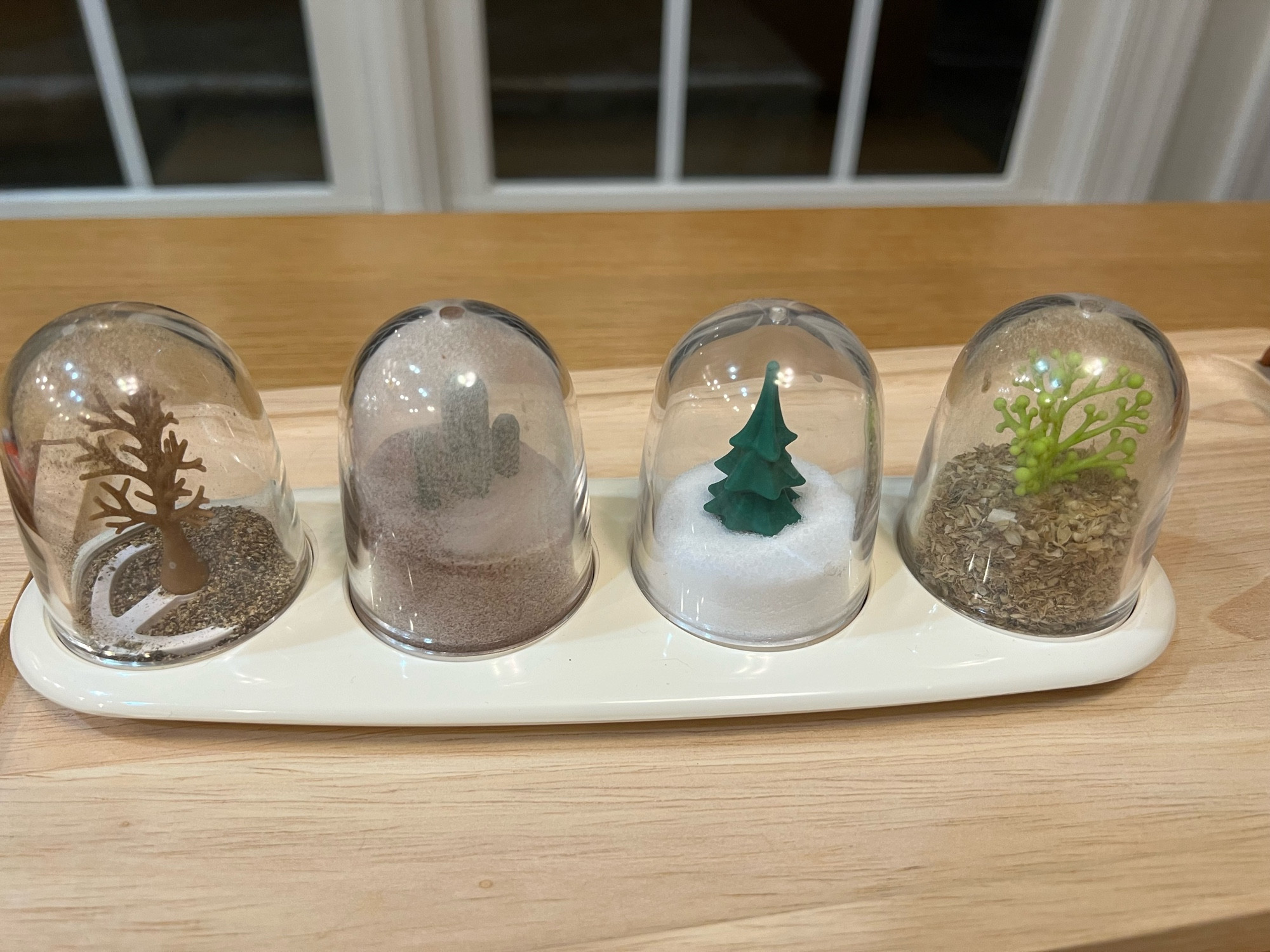 A condiment shaker set of four shakers, various trees in plastic bubbles. A brown tree with a small amount of black pepper, a green cactus submerged in cinnamon sugar, a green fir tree half buried in salt and a bright green spring tree in oregano.