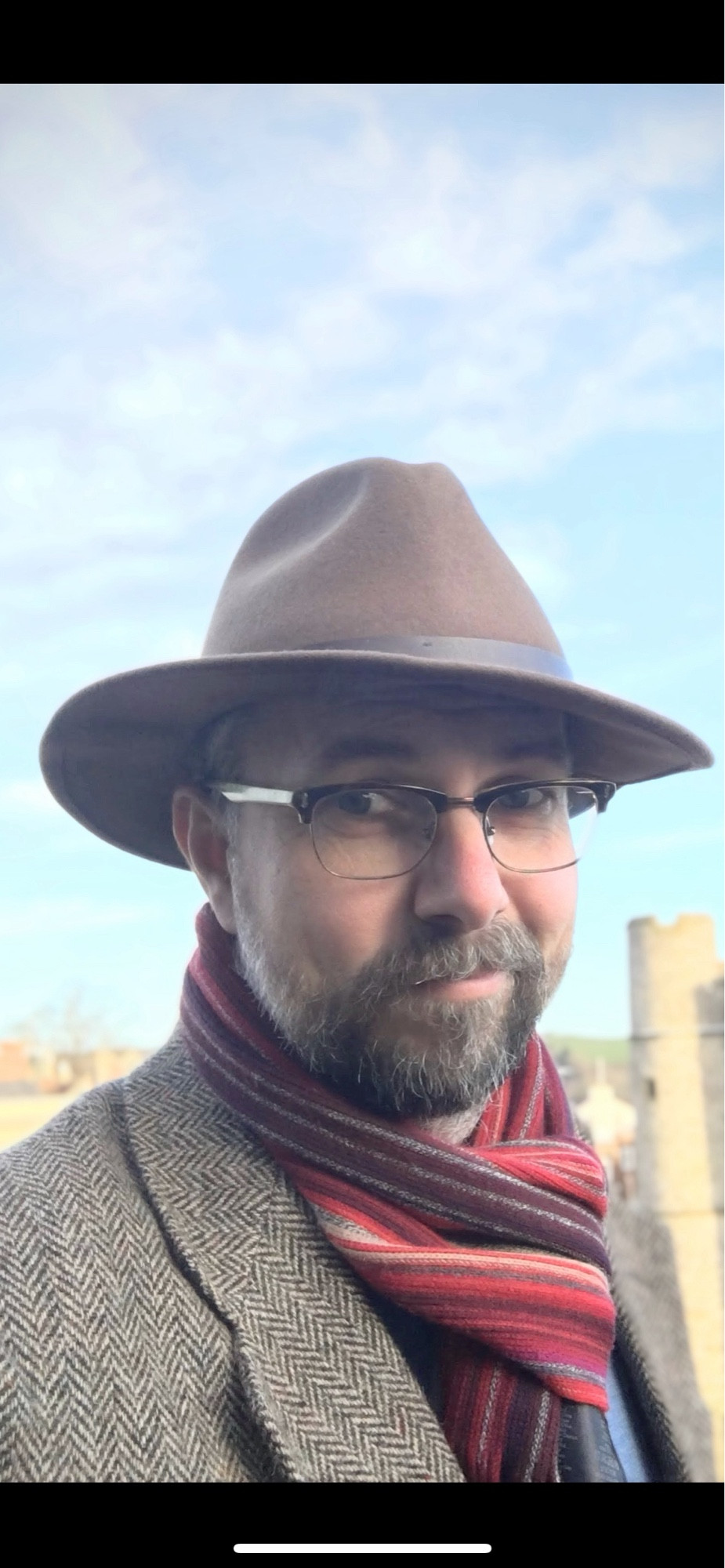 Ant wearing his fedora with a tweed jacket and a striped red and orange scarf on a sunny day at Lewes castle. Old photo with a lot less grey in the beard.