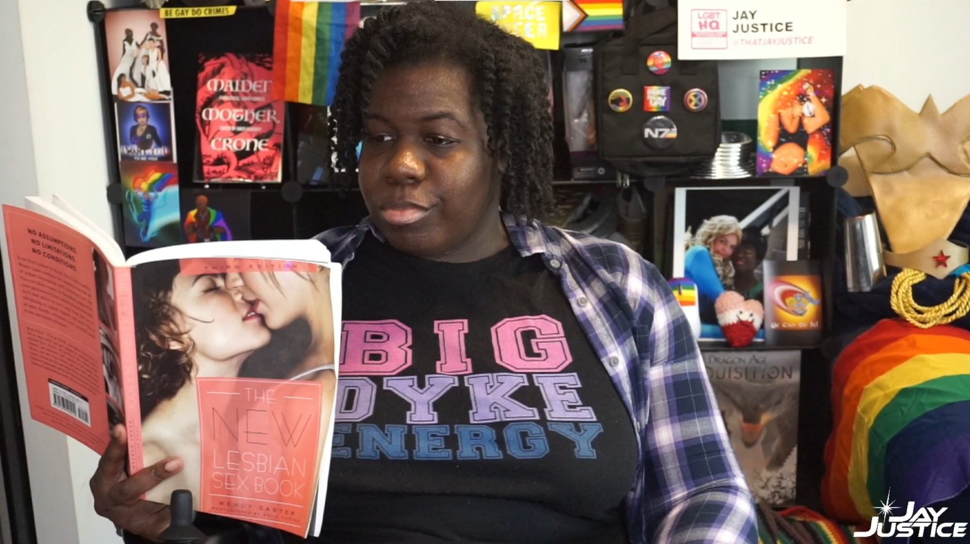 A dark-skinned Black woman with natural hair seated in a power wheelchair, wearing a t-shirt that says Big Dyke Energy, an open plaid button-down shirt, holding 'The NEW Lesbian Sex Book;, surrounded by rainbow flags, lesbian, trans, bisexual, pansexual, and queer artwork, images, pins, books and costume props created by and/or belonging to queer characters 