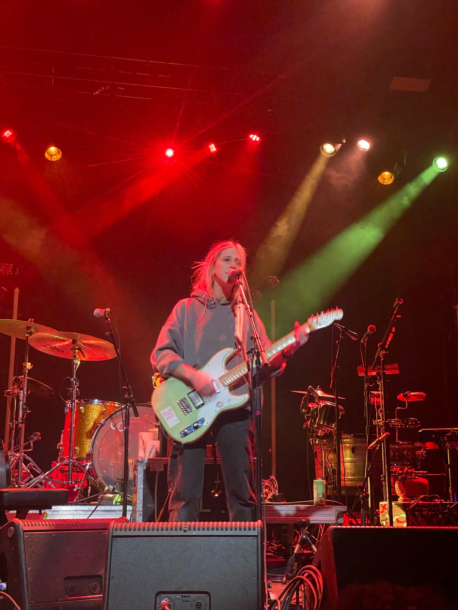 a blonde haired girl in a grey sweatshirt sings into a microphone while playing a mint green electric guitar. the stage behind her is filled with other instruments, illuminated in red light.