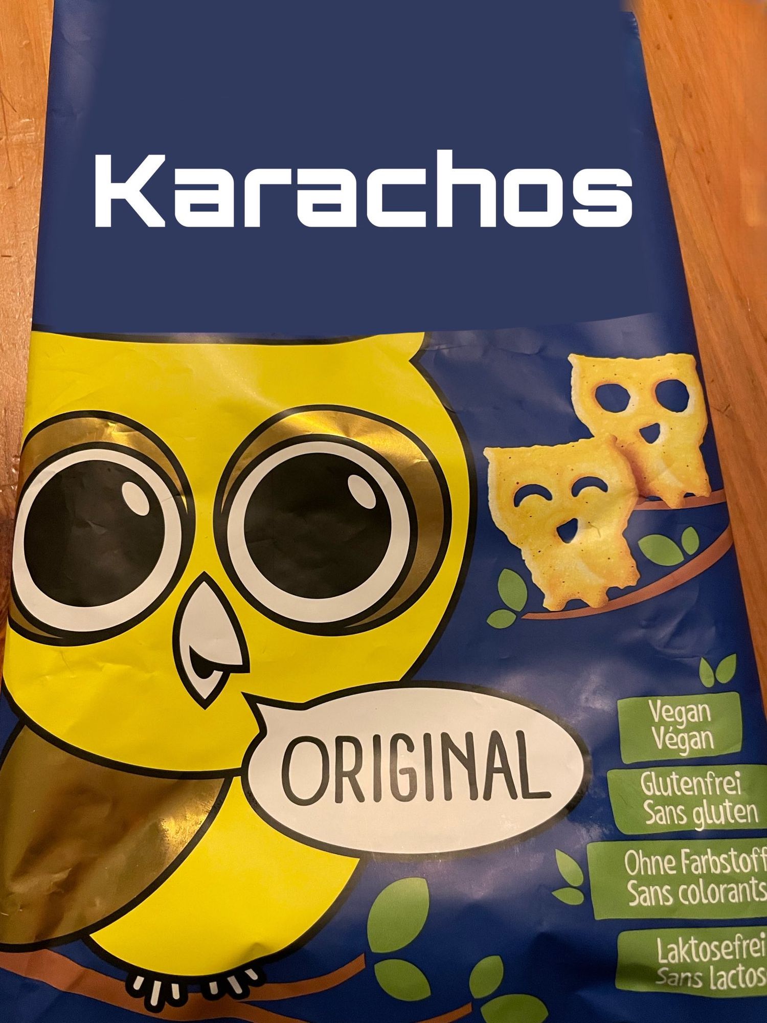 Das Bild zeigt eine bearbeitete Verpackung von Snacks der Marke "Karachos" die eigentlich Filou heisst und von der Marke Zweifel ist. Die Verpackung ist überwiegend in den Farben Blau und Gelb gestaltet. Im oberen Bereich der Verpackung steht der Markenname "Karachos" in großen weißen Buchstaben vor einem dunkelblauen Hintergrund.

Auf der Verpackung ist ein großes, cartoonartiges Bild einer gelben Eule mit großen, runden Augen zu sehen. Die Eule hält ein Schild mit der Aufschrift "ORIGINAL". Daneben sind zwei der Snacks abgebildet, die die Form kleiner Eulen haben.

Auf der rechten Seite der Verpackung befinden sich mehrere grüne Labels mit weißen Schriftzügen, die darauf hinweisen, dass die Snacks Vegan, Glutenfrei, Ohne Farbstoffe und Laktosefrei sind. 

Der Hintergrund der Verpackung ist dunkelblau mit grünen Blättern als Dekoration.