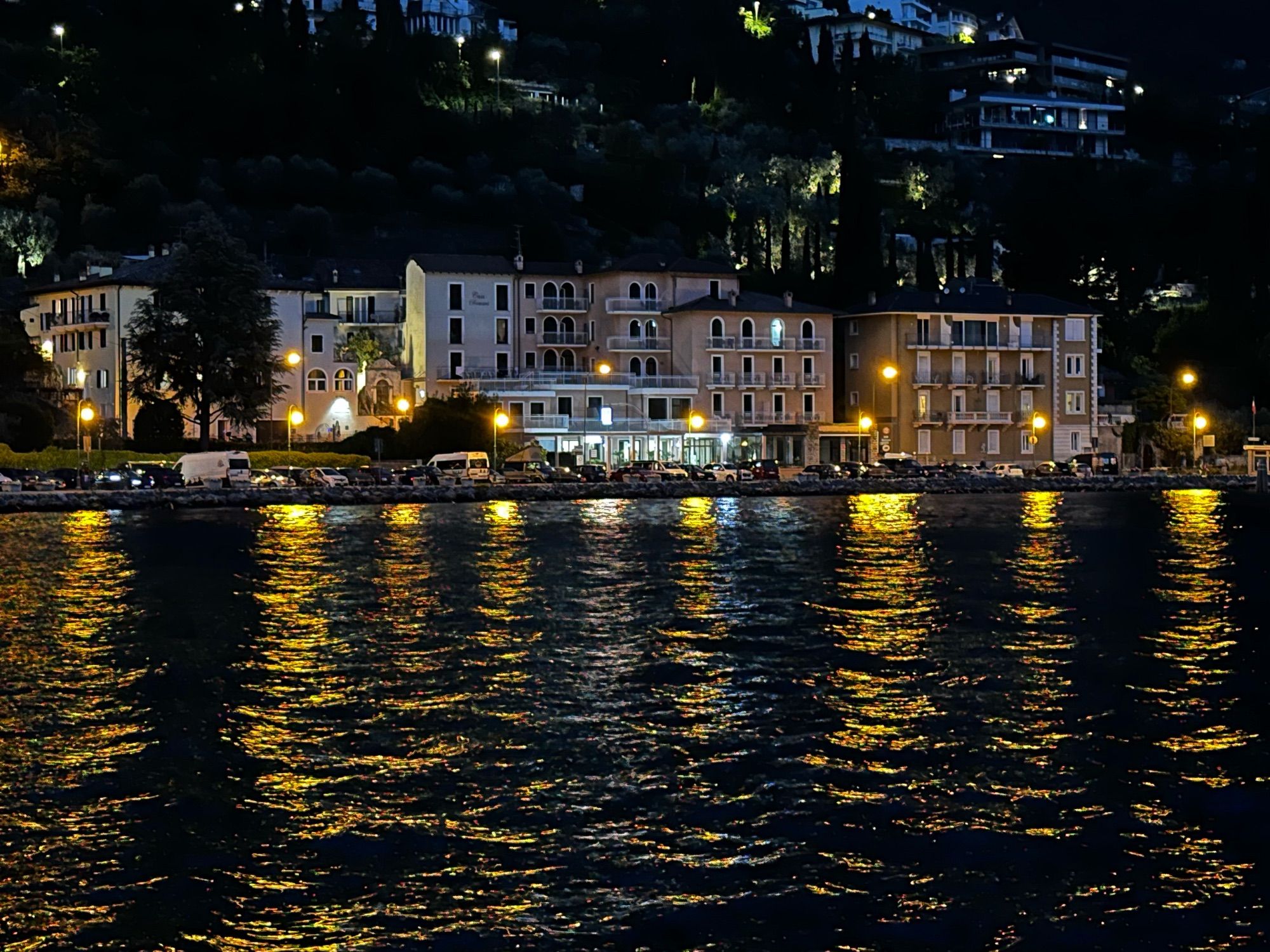 Nachtaufnahme einer Promenade mit beleuchteten Häusern, die Lichter spiegeln sich im See