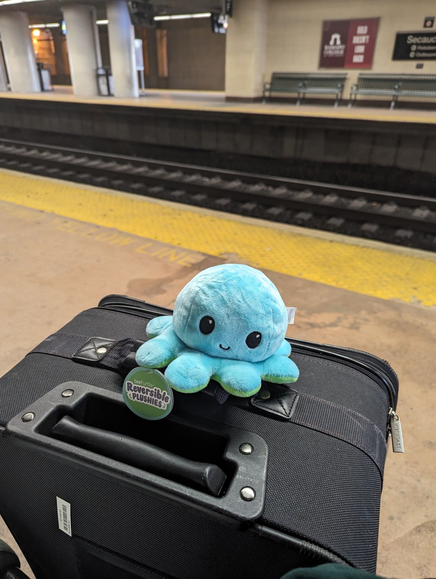 Blue plush octopus sitting on a suitcase next to some train tracks at a train station.