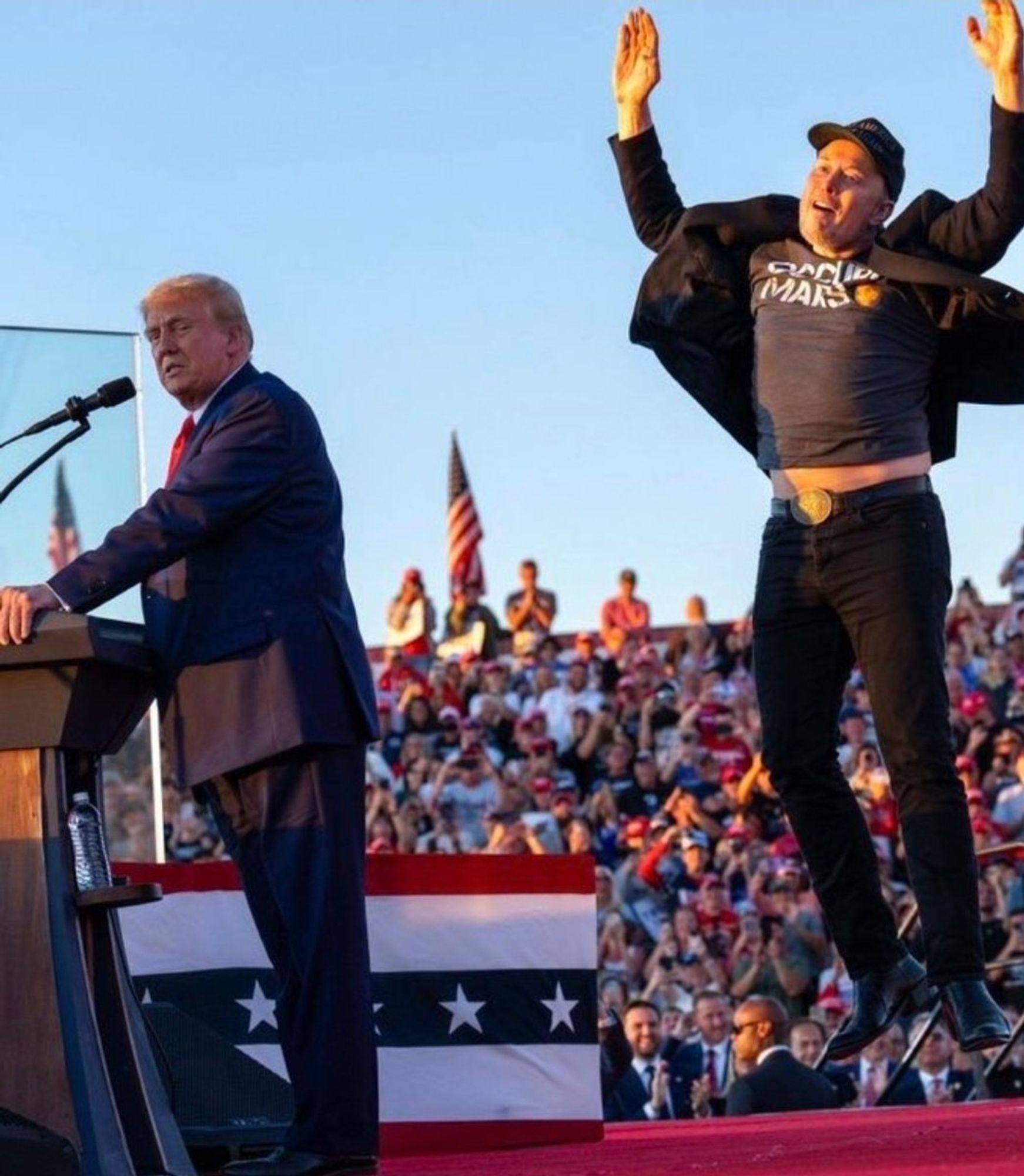 Elon Musk jumping behind an oblivious Donald Trump while on stage at a rally.