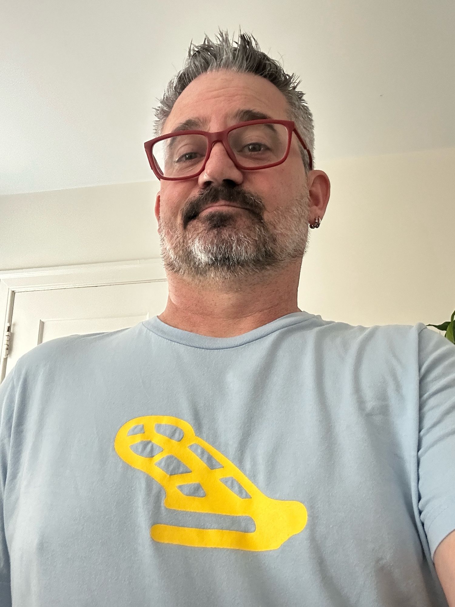 A selfie with “bear face” wearing a light blue t shirt with a yellow cobra chastity cage image on the front