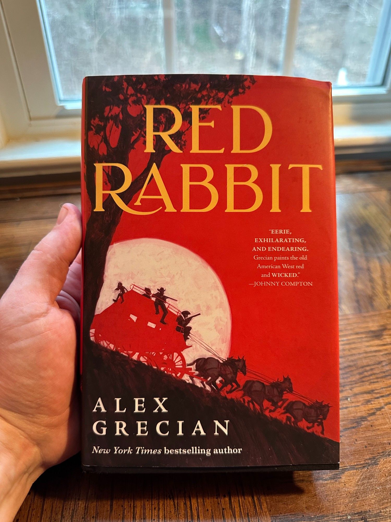 A hand holds a hardbound copy of RED RABBIT, by Alex Grecian above a wooden desk. The cover is mostly red, showing a stylized silhouette of a red coach and its team of horses racing down an incline. Four figures sit on the coach, and two hold long guns. A full moon is behind them.