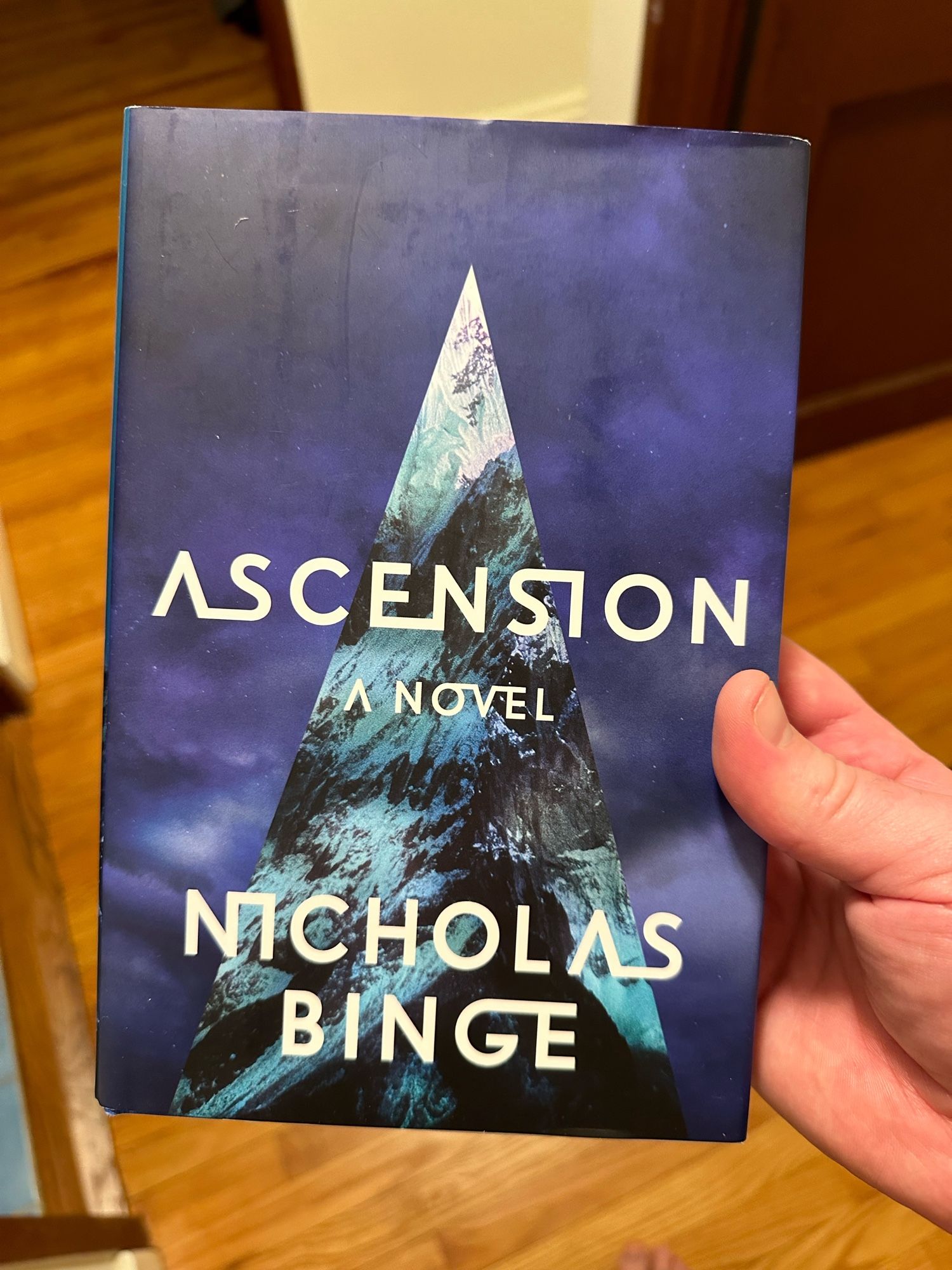 A hand hold a hardcover copy of Nicholas Binge’s ASCENSION. The cover is blue, reminiscent of steel storm clouds, and a mottled teal and dark blue/black triangle rises from the bottom of the cover.