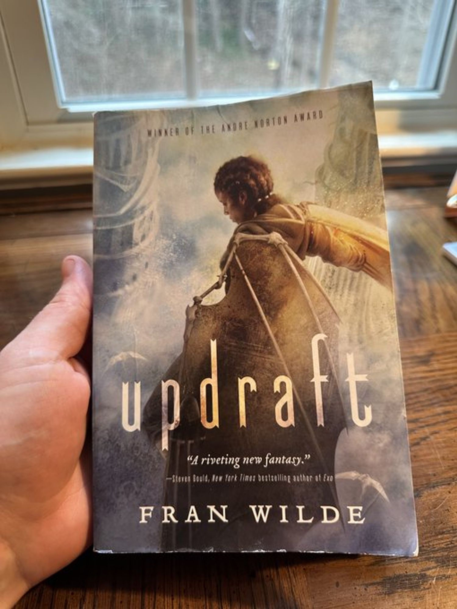 A hand holds a paperback copy of UPDRAFT, by Fran Wilde above a wooden desk. A young woman dressed in a set of fabric and bone wings perches on an indistinct edge, as if about to leap into space. In the distance, ornate bone towers rise from the clouds.