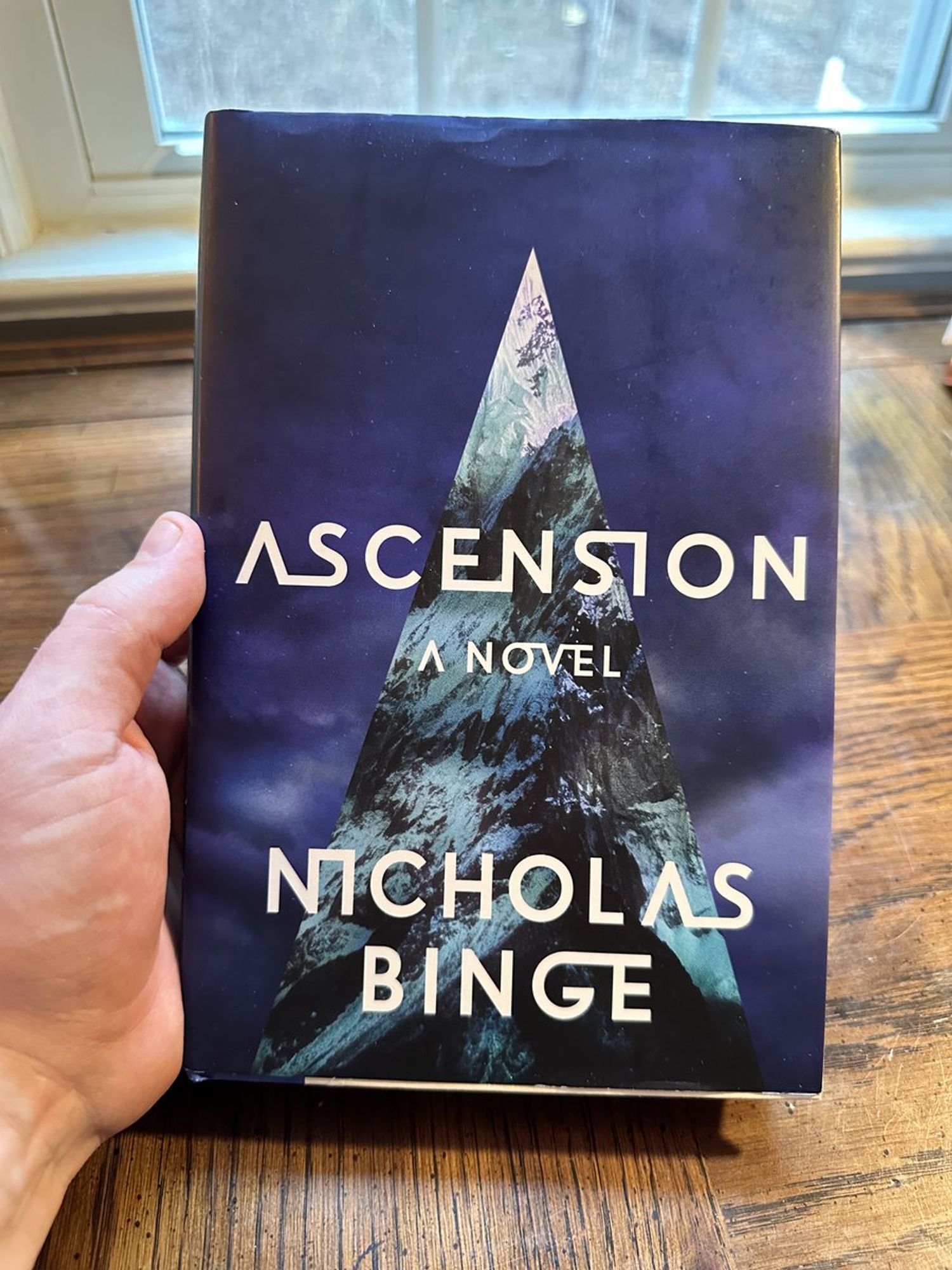 A hand holds a hardbound copy of ASCENSION by Nicholas Binge over a wooden desk. The cover is a mottled deep blue & purple, suggesting storm clouds. A spire of teal and black rises from the bottom in a sharp triangle.