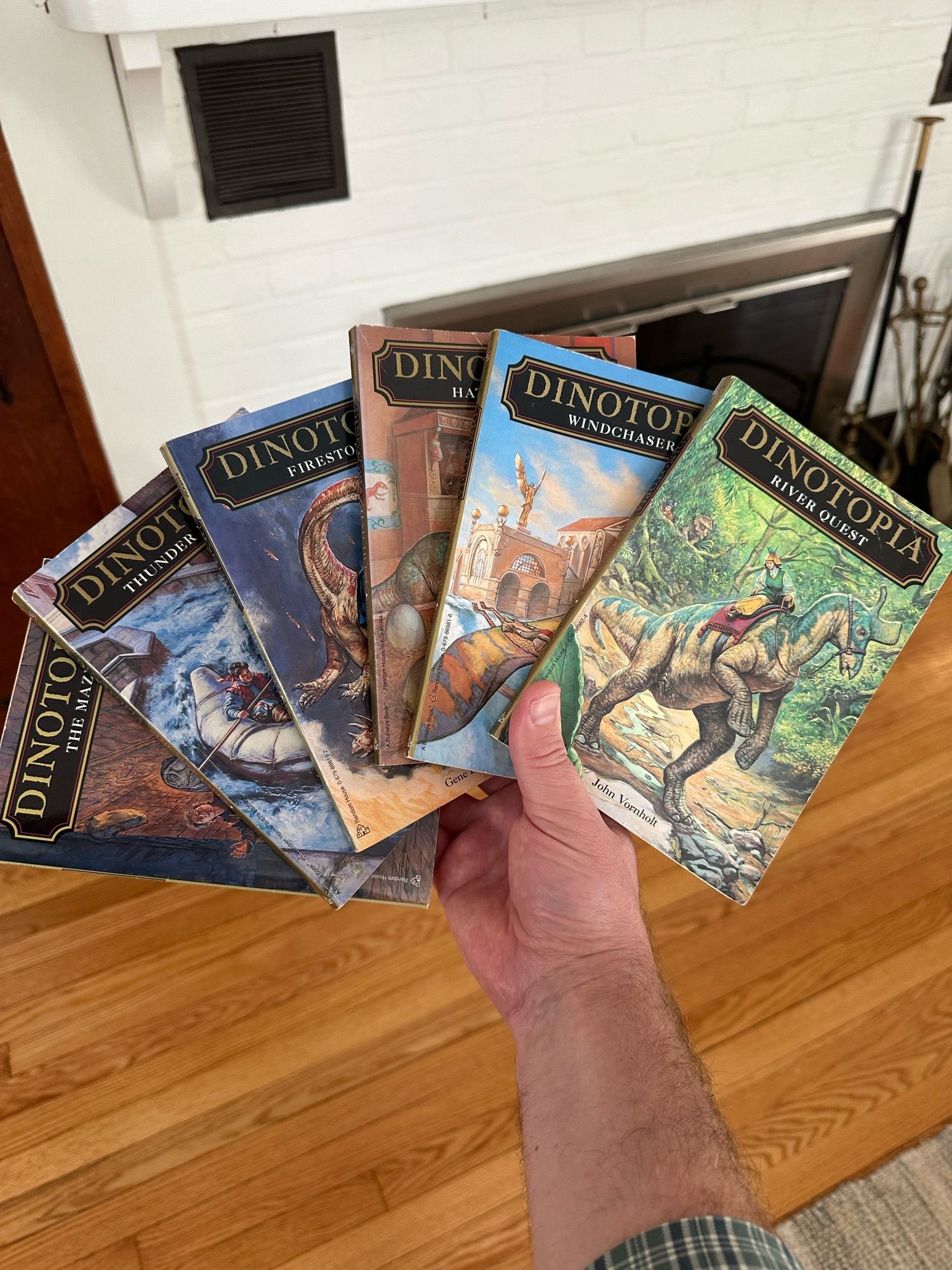 A hand holds out six slim paperback books like a hand of cards, each a Dinotopia novel with beautifully painted covers of dinosaurs and humans coexisting. A white painted fireplace is in the background.