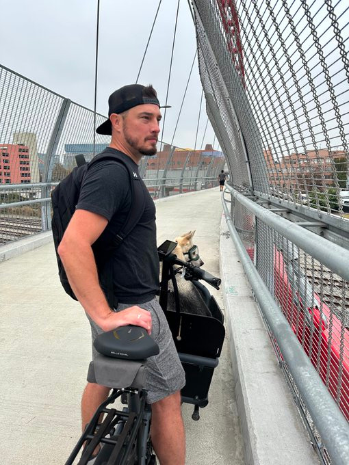 Man wearing shorts, black t-shirt, and backwards black baseball hat on a bike on a bridge. A dog is in the front of his bike.