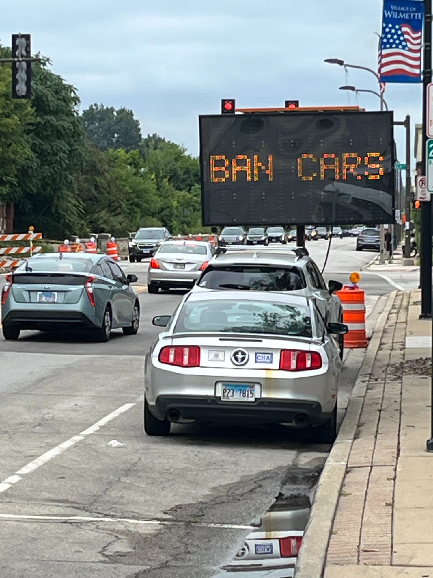 Road with orange construction barrels on it and a large variable message sign that says BAN CARS in digital letters. A silver mustang is parked in the foreground.