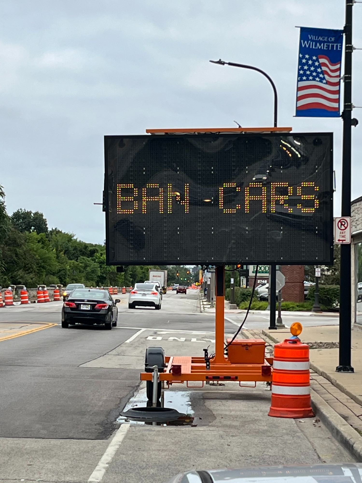 Road with orange construction barrels on it and a large variable message sign that says BAN CARS in digital letters.