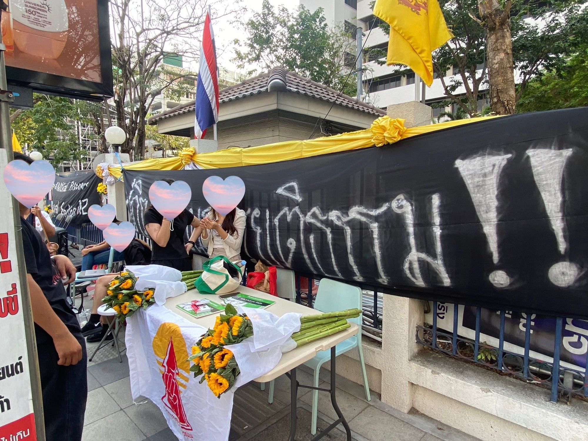 Thai activists preparing for discussion & activities outside Ratchada Criminal Court. 
Banner behind them reads #บีบแตรไม่ใช่อาชญากรรม Honking horn is not a crime
On the table in front of them are sunflowers and the flag of แนวร่วมธรรมศาสตร์และการชุมนุม (UFTD) / Thammasat UFTD