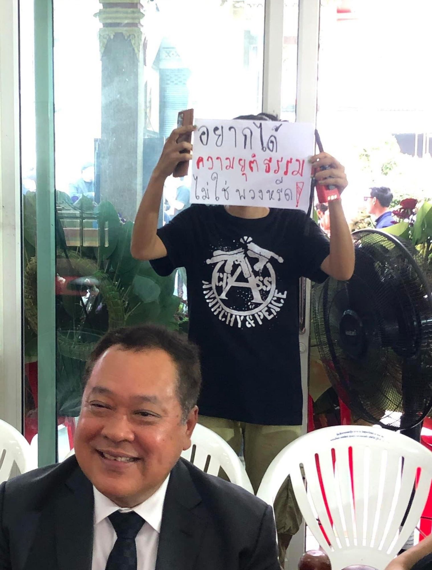 Activist holds up sign behind Thai justice minister which reads 

อยากได้ความยุติธรรม ไม่ใช่พวงหรีด

We want justice not a wreath

Activists t-shirt reads anarchy & peace.