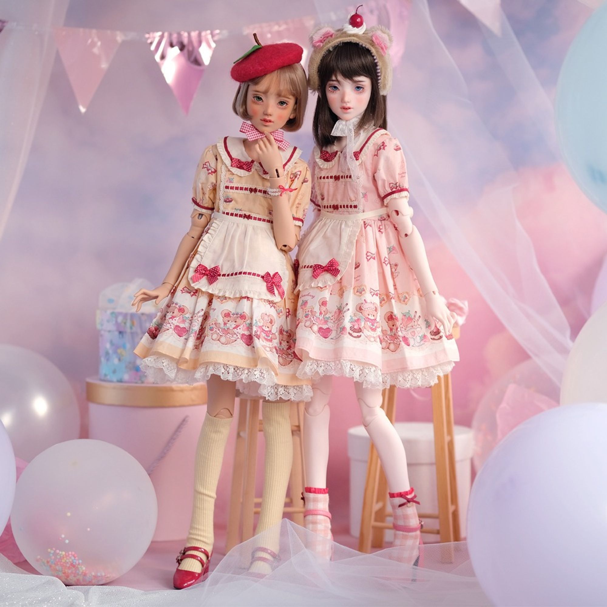 Two dolla in pastel themed dresses pose standing for a photo. They stand in a pastel pink-purple party decoration.