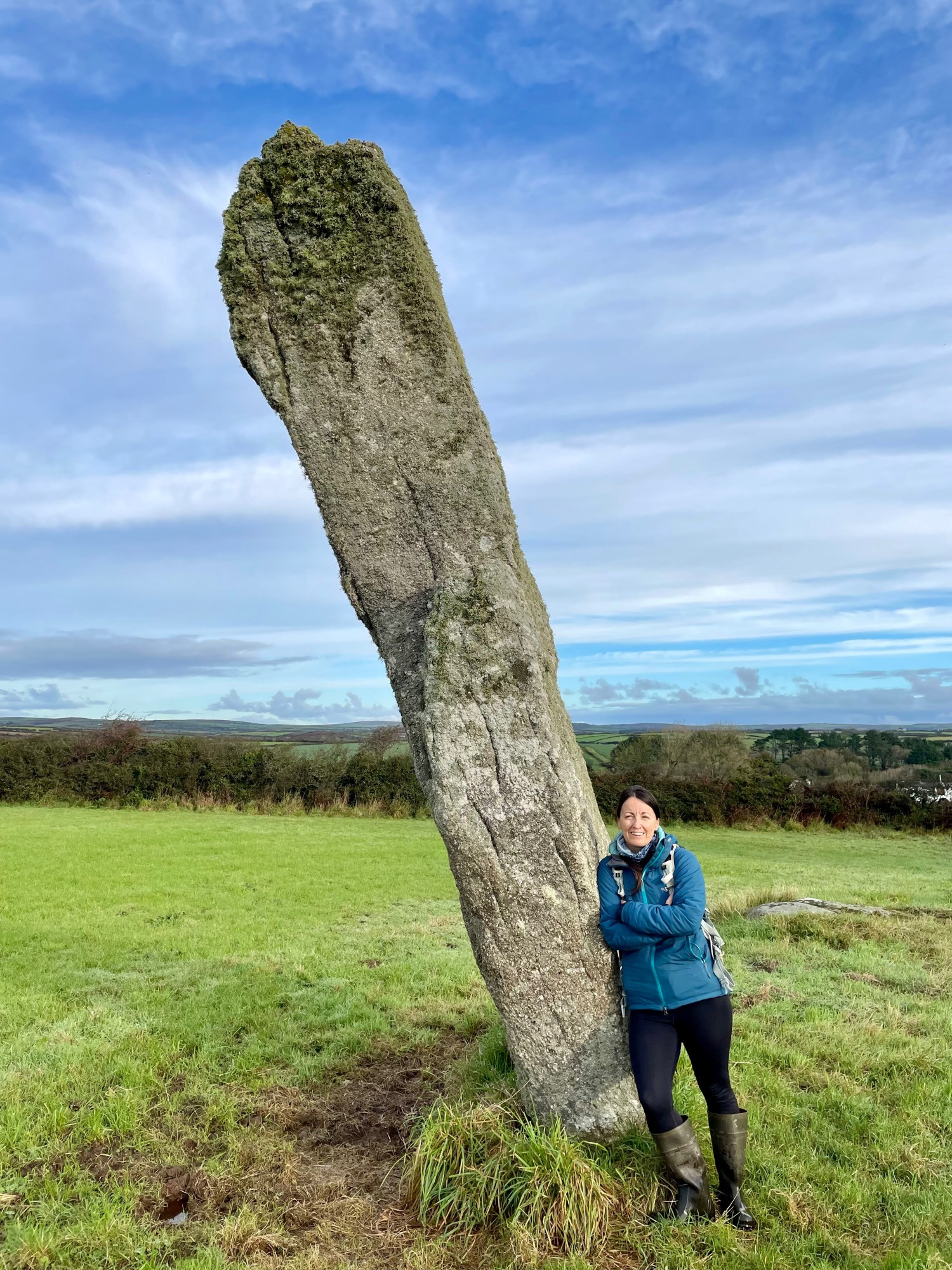 A dark haired woman (ie me) in a blue coat leaning against an ancient standing stone in a field.