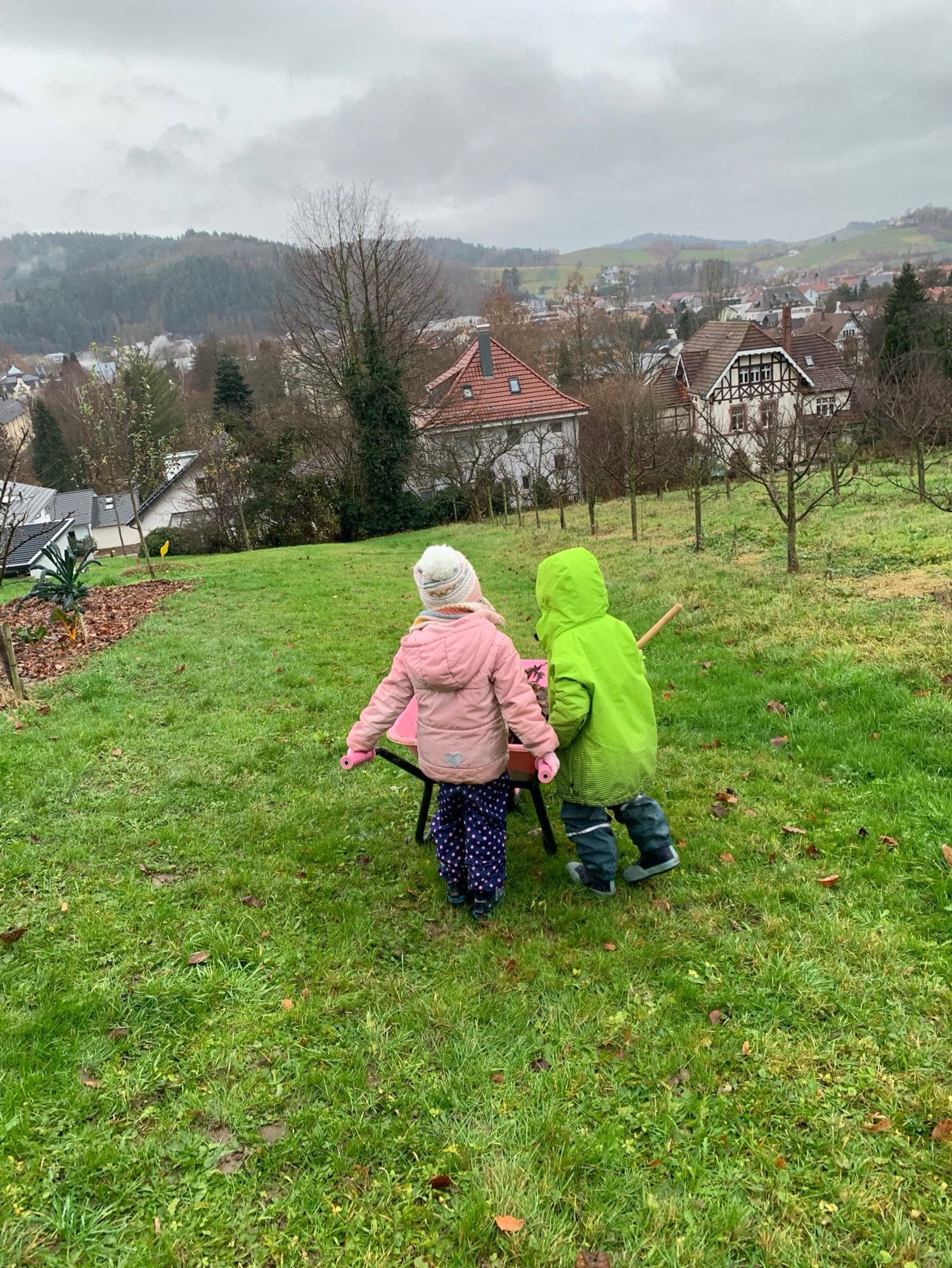 Zwei Kinder fahren einen Schubkarren voll Laub den Hang hinunter.