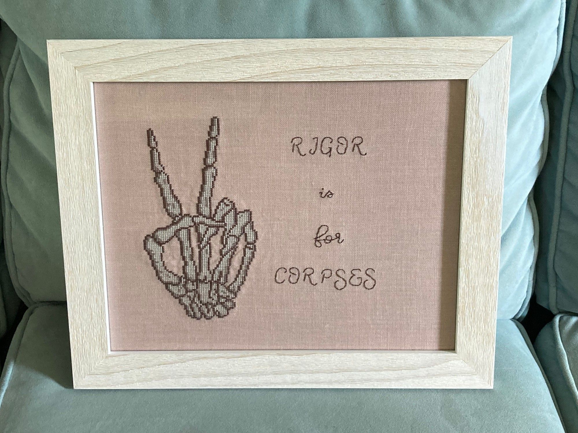 Cross stitch piece on light pink fabric in sepia tones. Shows a skeleton hand giving the peace sign next to the text "RIGOR is for CORPSES."