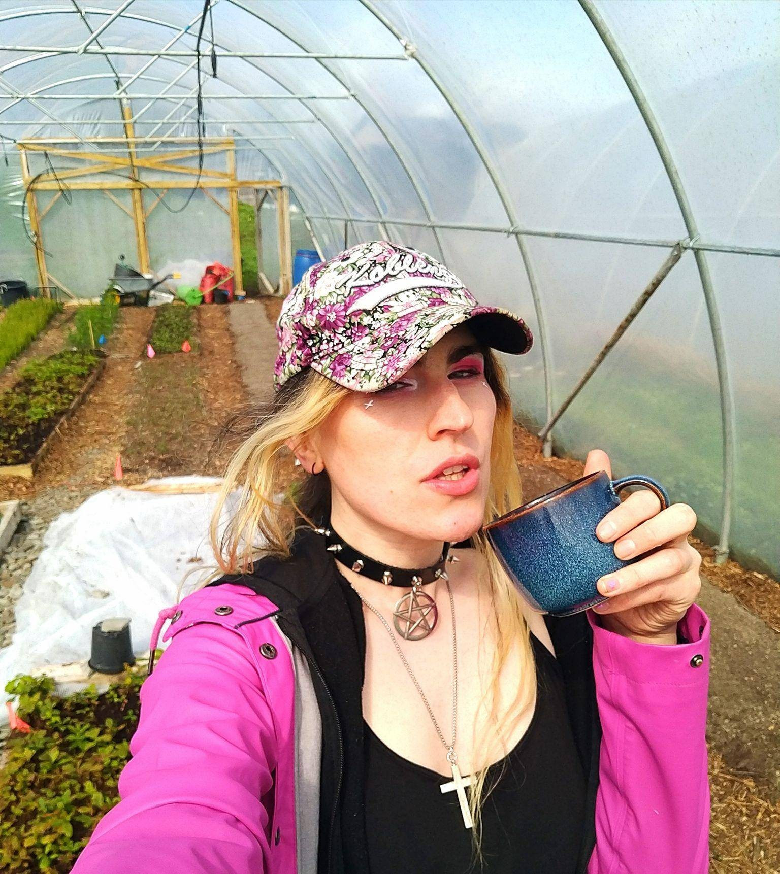 Selfie of white goth girthing in a garden poly tunnel. She is wearing a pink jacket, cap, a crucifix chain and a pentagram collar. She is holding a cup of tea and behind her are rows of little saplings in the poly tunnel.