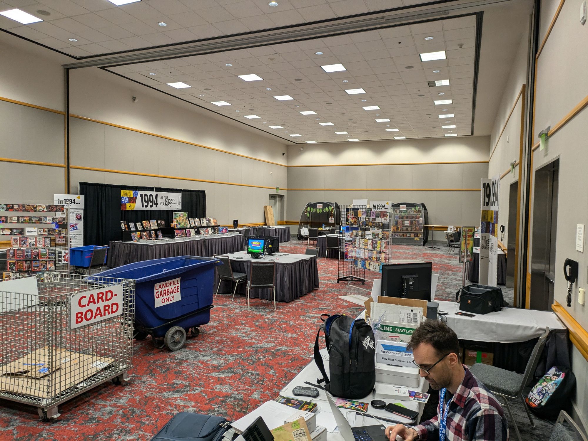 Under-construction shot of the VGHF museum room at Portland Retro Gaming Expo 2024. Frank is visible, bottom-right, doing something on his laptop.