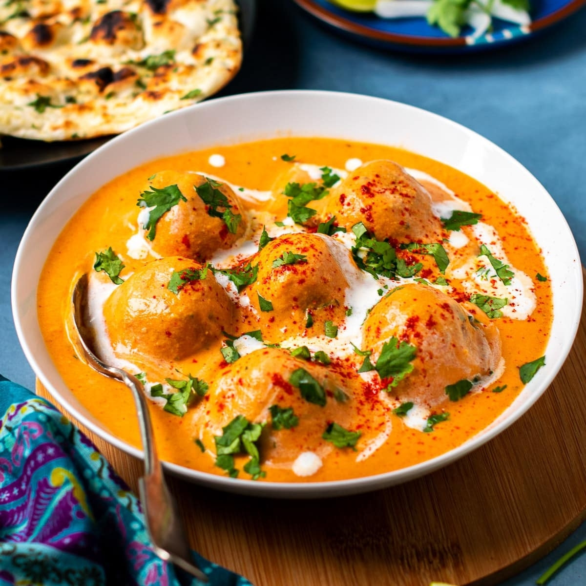 In the foreground: A spoon & bowl of fried balls of paneer (Indian cheese) in a rich and creamy orange-colored "gravy" made with sweet onions and tomatoes. Drizzled with cream, dash of red powder, and sprinkles of coriander.  In the background: Some fluffy garlic naan on a plate