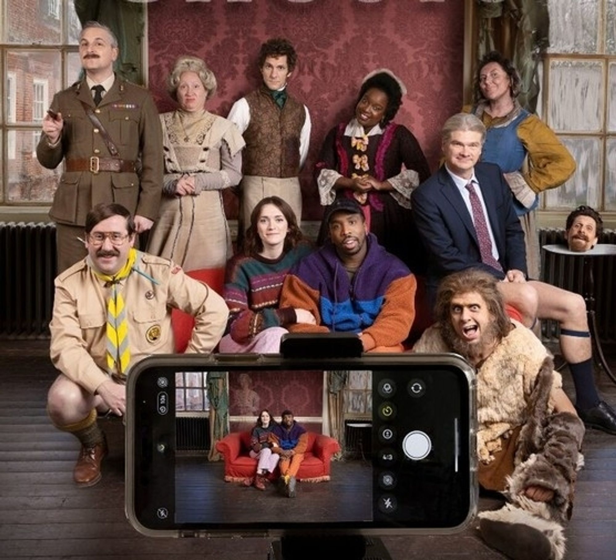 BBC ghosts. A young married couple sit on a sofa, flanked by 9 ghosts in different period costumes. They are all posing for a photo being taken on a phone. On the phone screen we can only see the young couple and, very faintly, Fanny the edwardian ghost