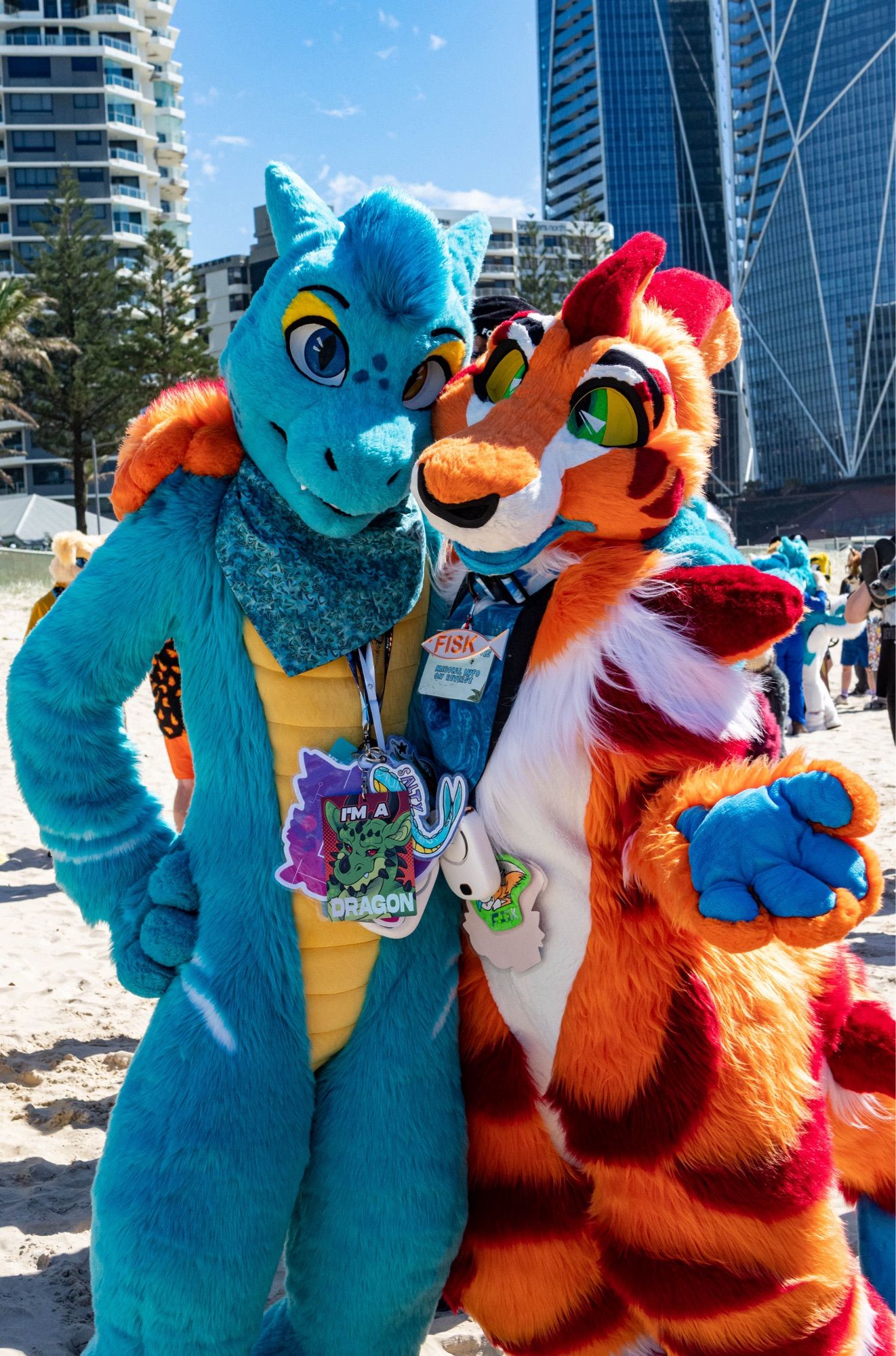 A blue aquatic dragon fursuiter and a orange shark-hound fursuiter standing together Posing for a photo on the beach in QLD, Australia at FurDu 2024