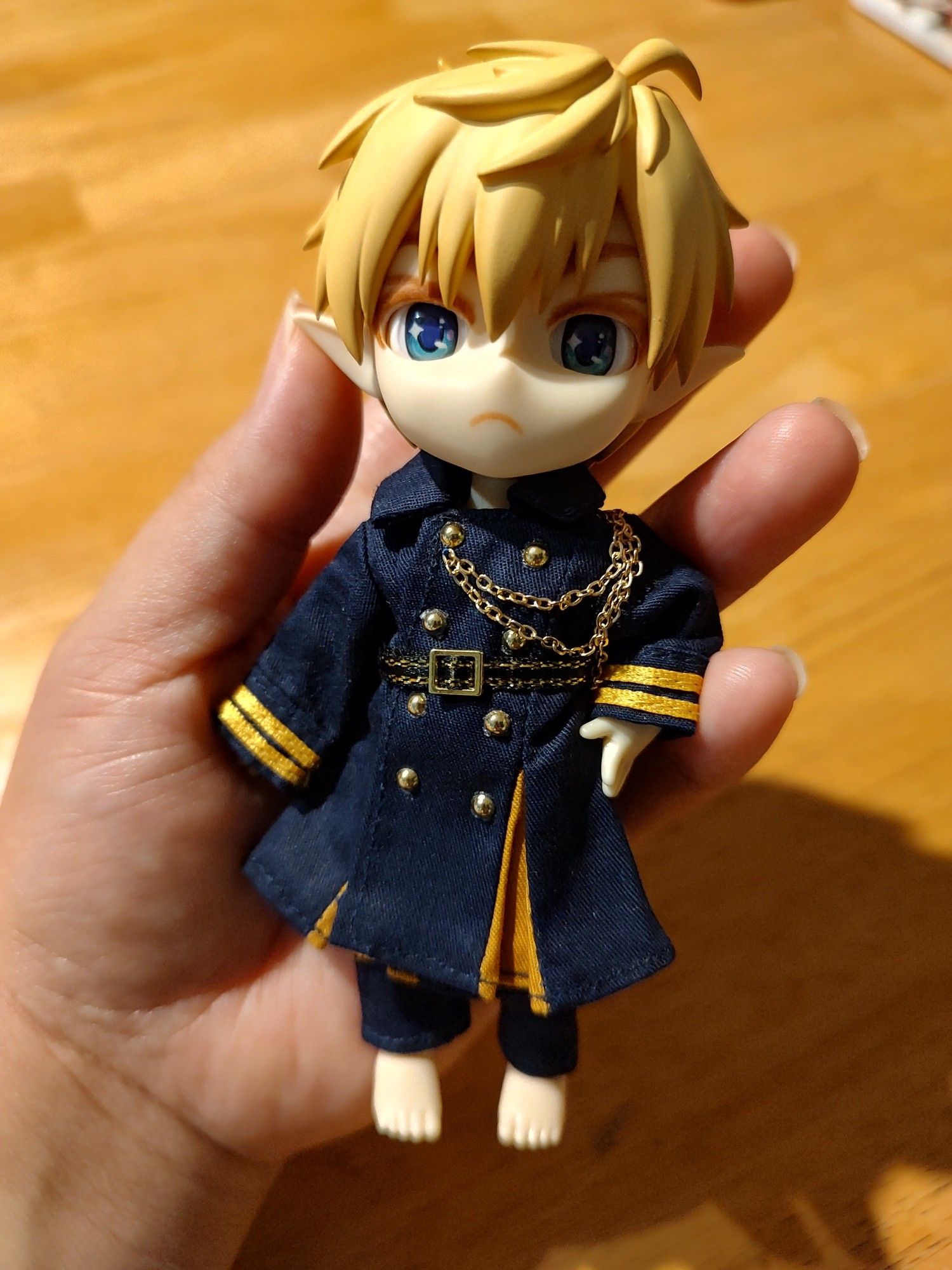 A customized 1/12 scale doll resting in the palm of my hand. He has plastic molded blond hair, plastic matte blue eyes, elf ears, and a grumpy expression. He is wearing a navy blue military style outfit with gold accents, and no shoes.