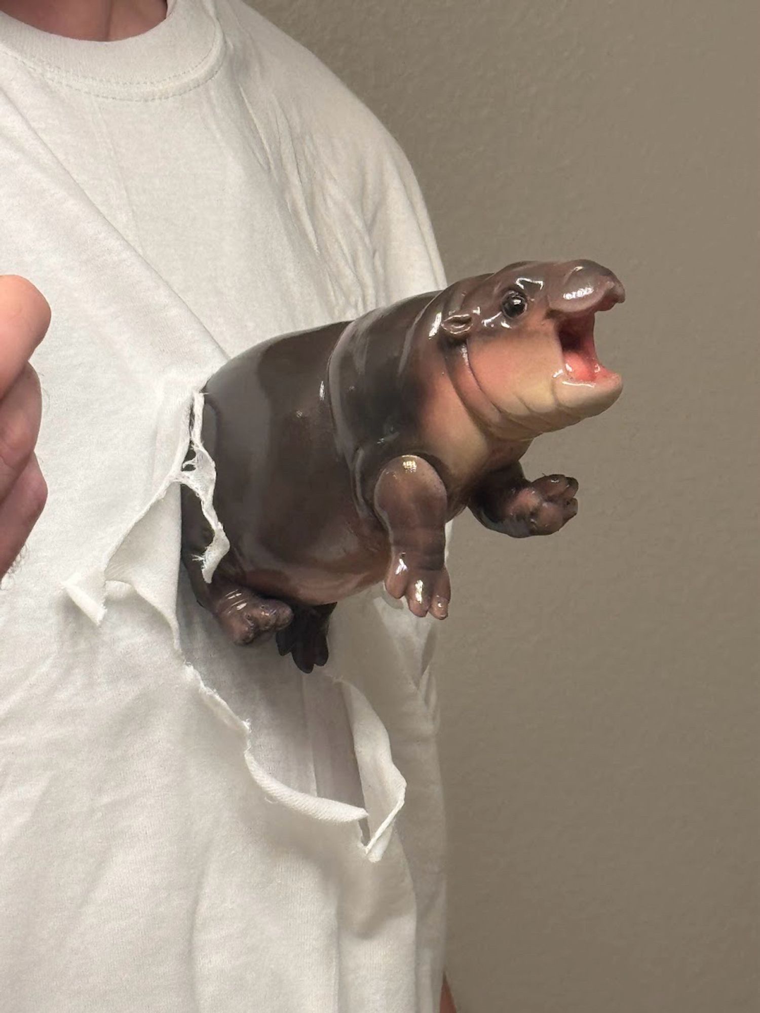 A small painted baby hippo statue ripping its way out of a plain white t-shirt on a presumably male model