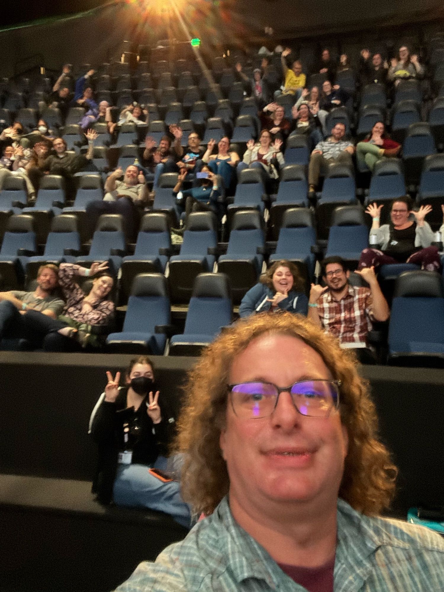 A man in a blue shirt with long curly hair smiling in a selfie with a half-full auditorium behind him