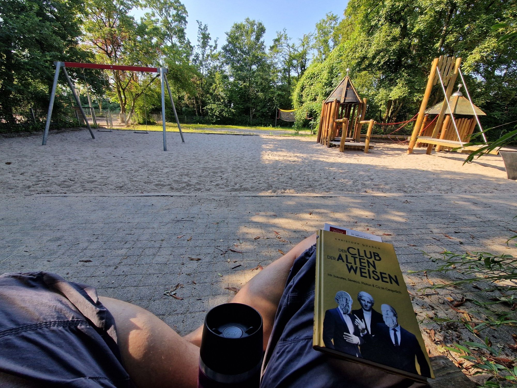 Blick auf einen Spielplatz, meine Beine im Vordergrund im Schneidersitz, der Kaffeebecher und ein Buch. Es ist noch schön schattig.