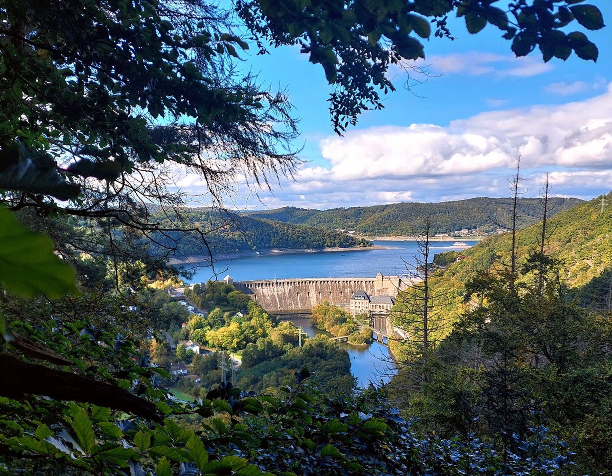 Blick von vorn oben auf die Staumauer des Edersees, gerahmt von Bäumen und Blätterwerk. Vor der Staumauer, das Ablaufbecken von Hemfurth, hinter der Staumauer der Edersee gut gefüllt, außerdem bewaldete Berglandschaft. Auf dem See ist ein AUsflugsschiff zu erkennen.