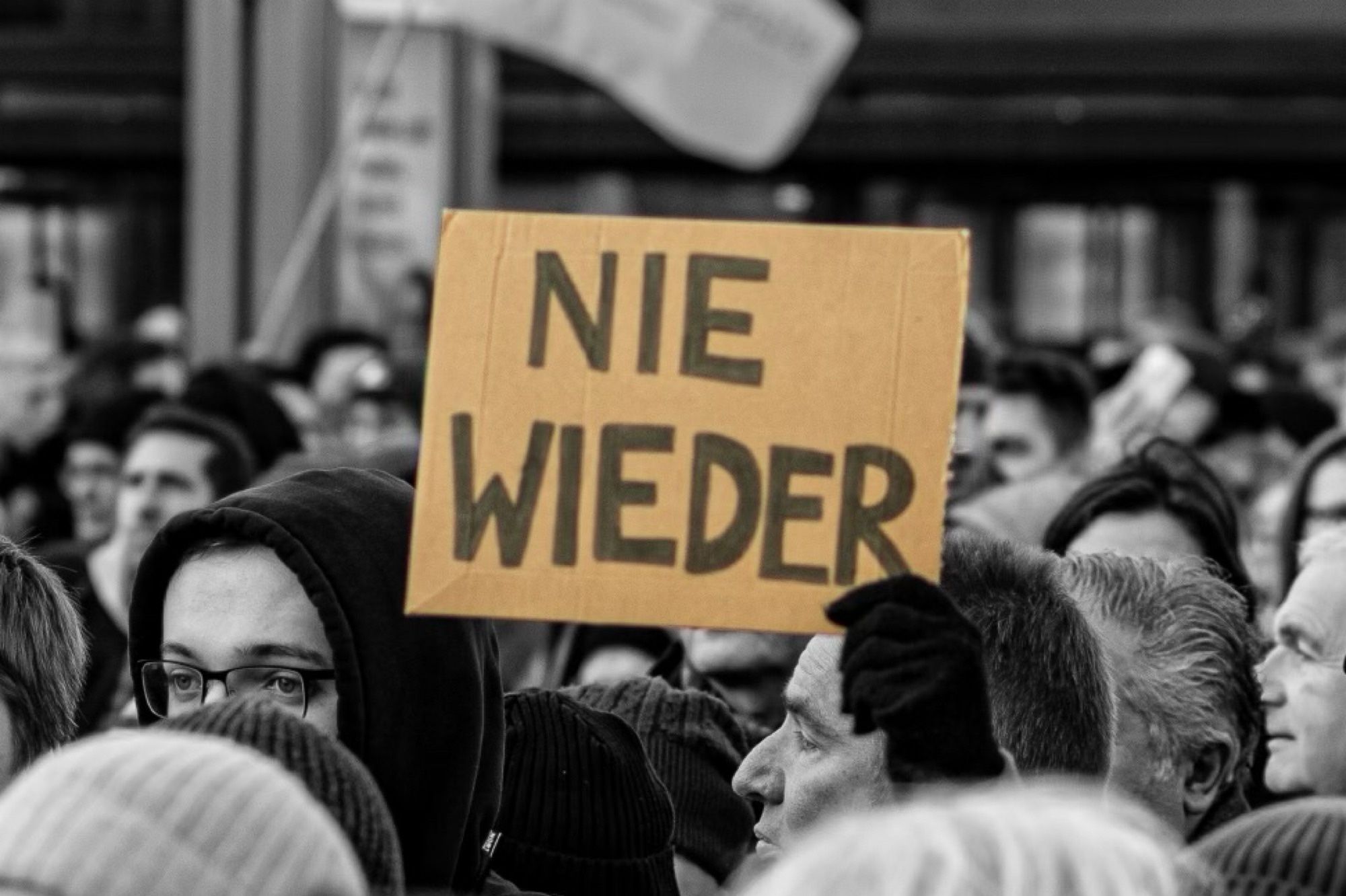 Ein SW-Bild von einer Demonstration. In der Mitte zwischen den Köpfen ein Schild mit der Schrift: Nie Wieder. ©️Sontje Neander