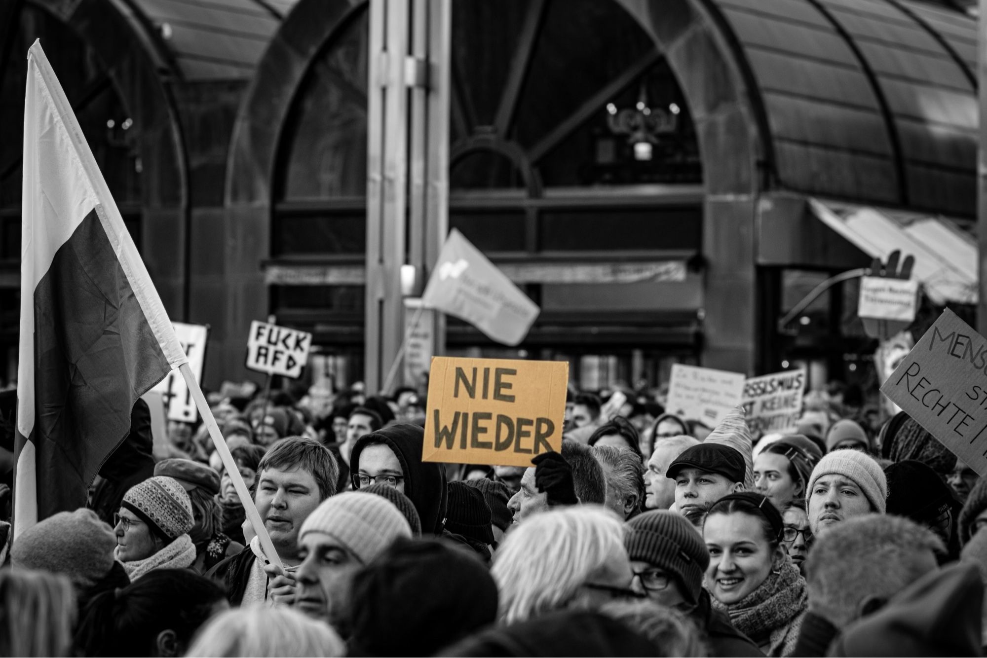 Eine Demo mit vielen Menschen in Schwarzweiß, dazwischen ein Schild mit dem Text „Nie wieder“.