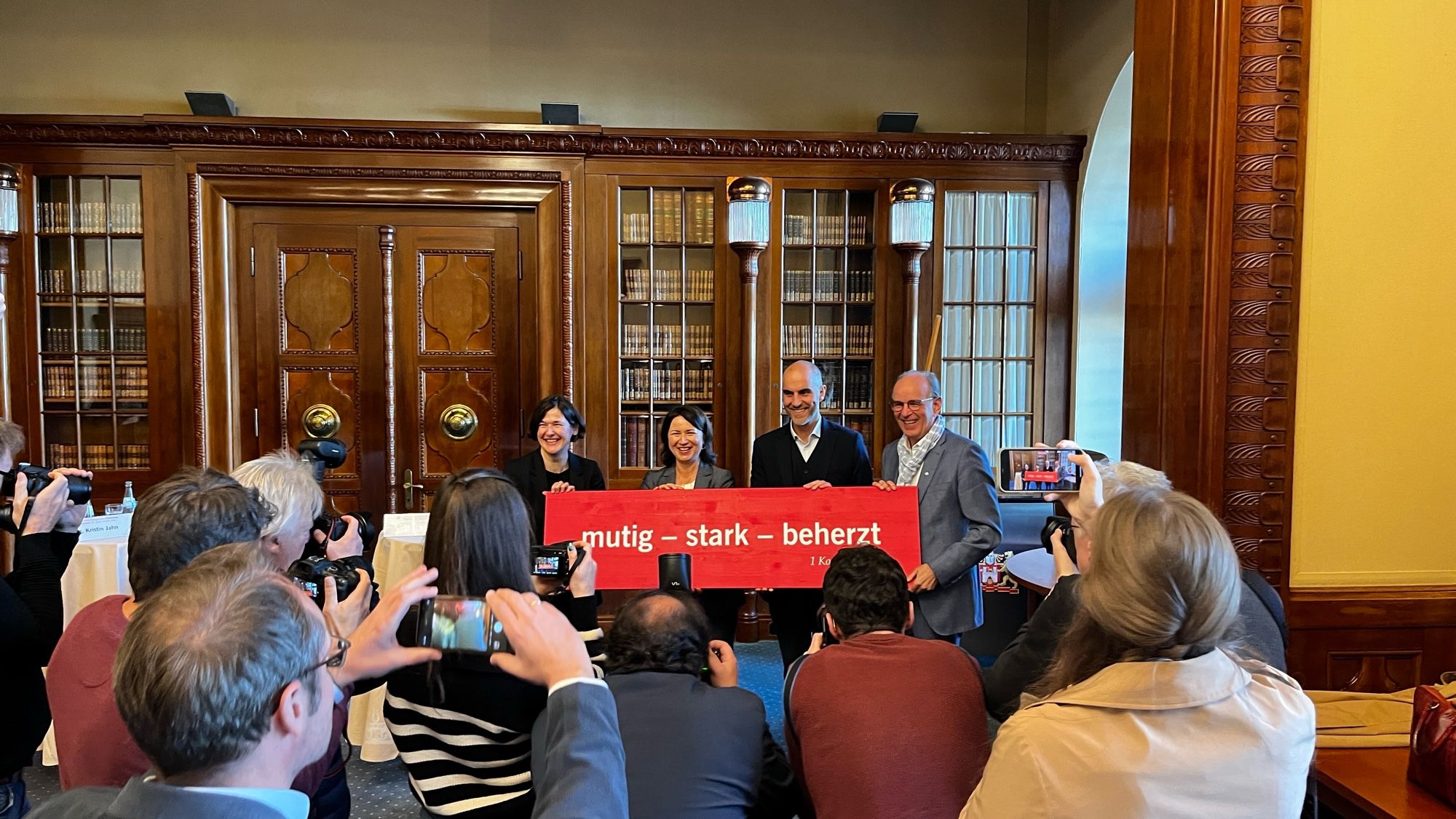 Vor einer Bibliothek stehen vier Personen, sie halten eine rote Tafel mit weißer Schrift: „Mutig-stark-beherzt“. Vor den Personen stehen viele Menschen mit Fotoapparaten.