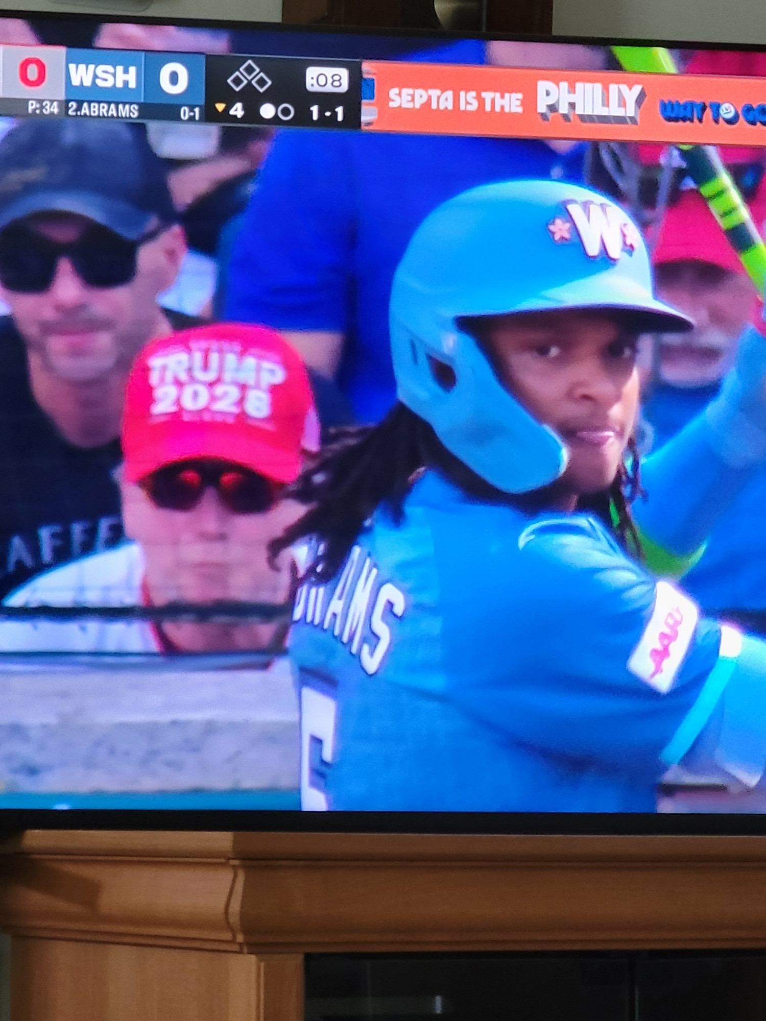 Washington Nationals fan in the front row behind the plate at a baseball game, wearing a red "TRUMP 2028" hat