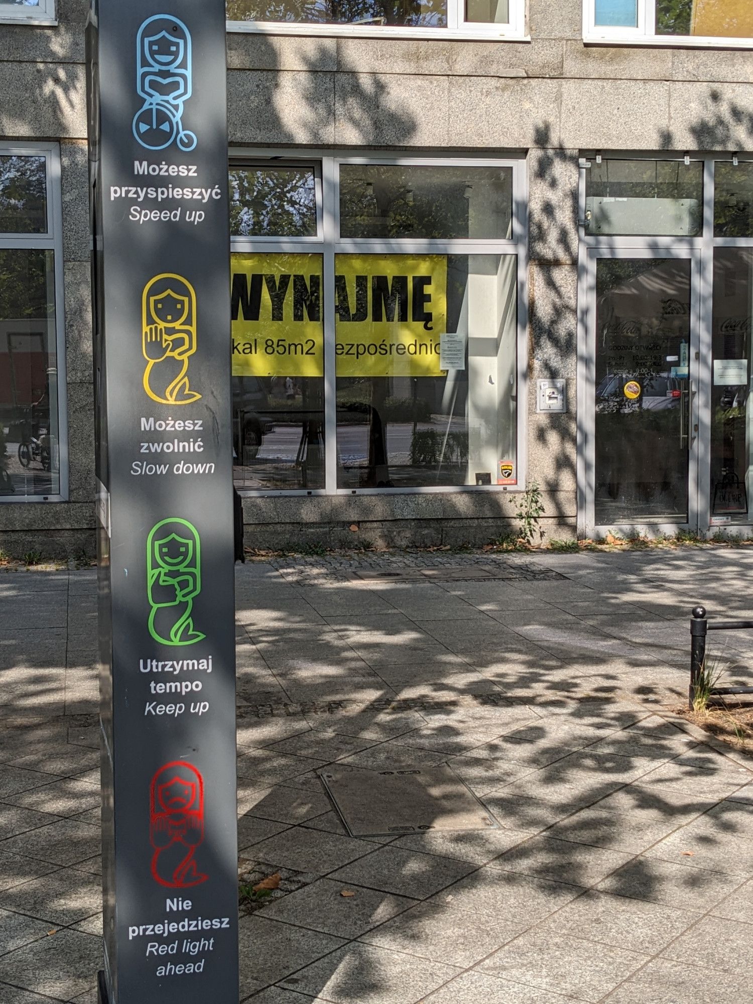 On a column signpost are 4 mermaids. The blue one rides a bike, with key text "Możesz prsyspieszyć. Speed up." The yellow one holds up a hand. "Możesz zwolnić. Slow down." The smiling green mermaid gives a thumbs up. "Utrzymaj tempo. Keep up." The frowning red mermaid warns with both hands: "Nie przejedziesz. Red light ahead."