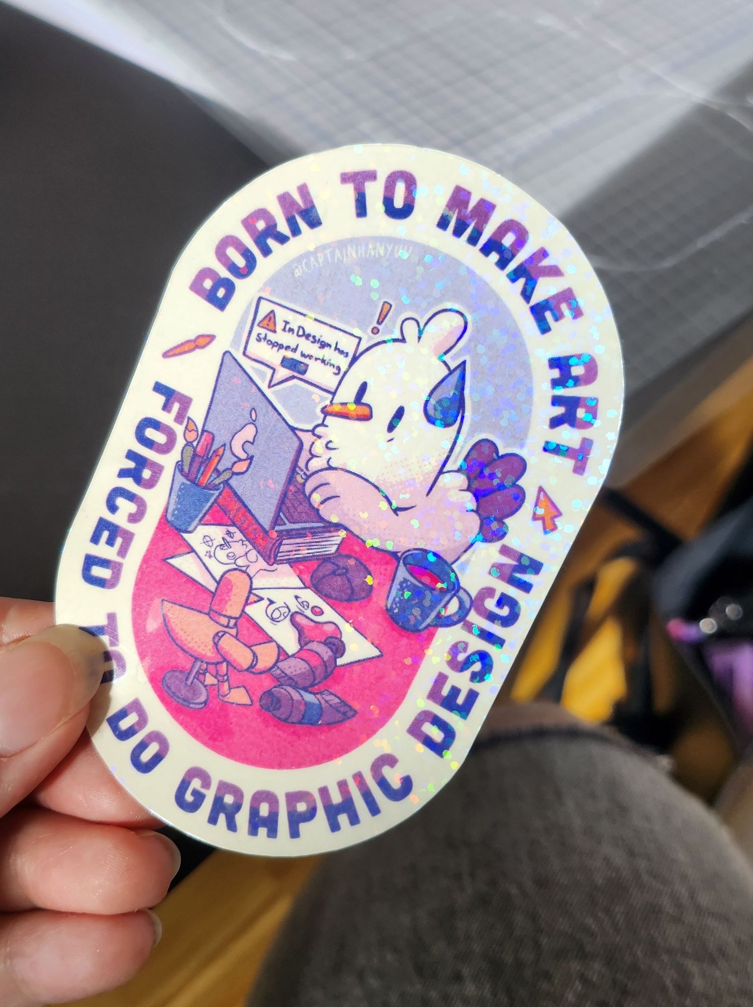 Photo of a glitter sticker. It reads "Born to make Art, forced to do graphic design". It's a seagull on a laptop surrounded by art supplies. InDesign has stopped working...