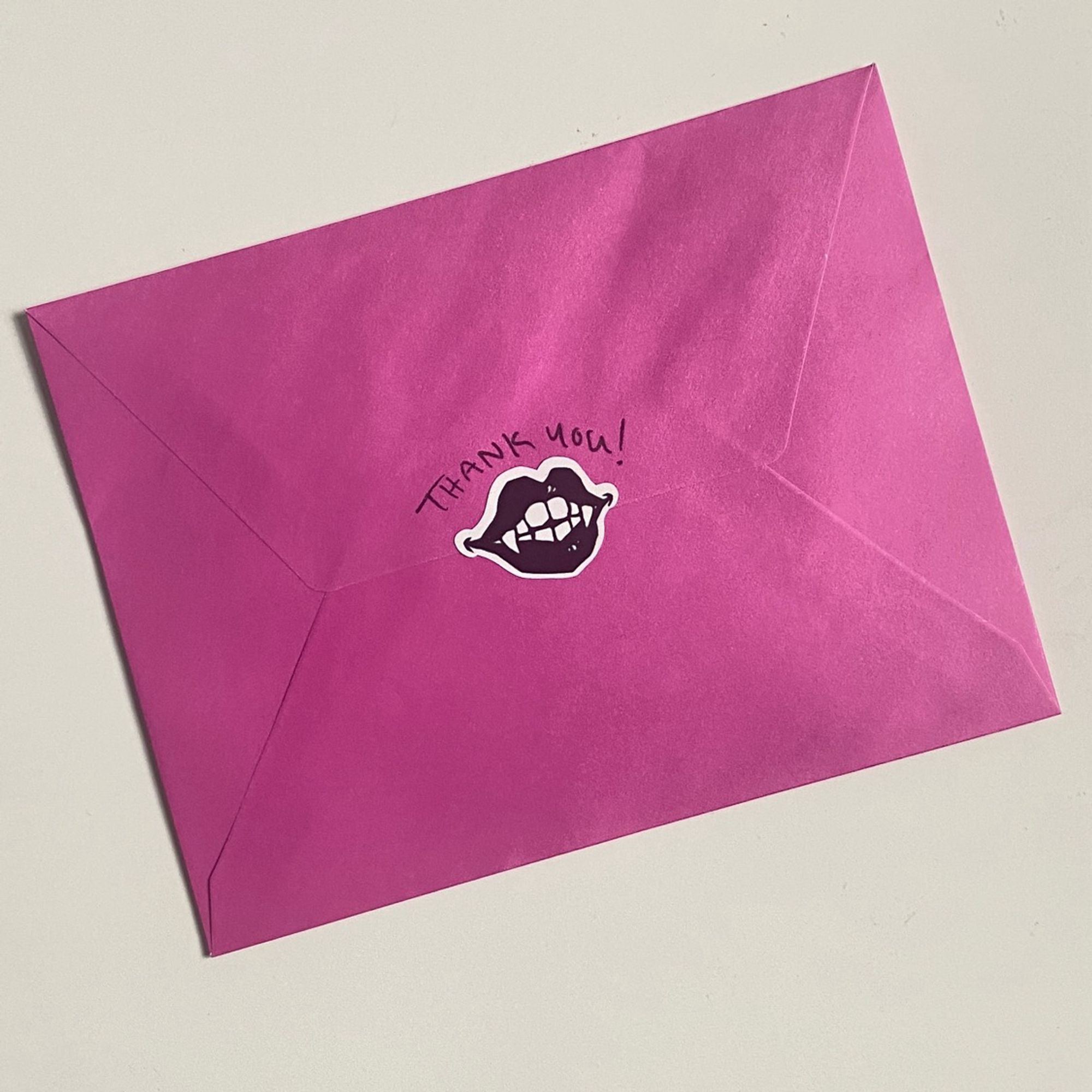 An example order from Live Flashy or Die. The back of a hot pink letter envelope with a small vampire smile and thank you!