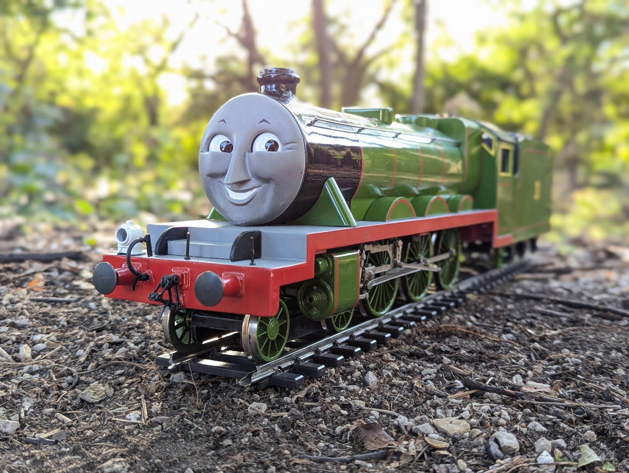 Henry the Green Engine as a Gauge 1 replica sitting on a piece of track in the middle of the woods. The afternoon sunlight shines through the trees and reflects beautifully off of his boiler