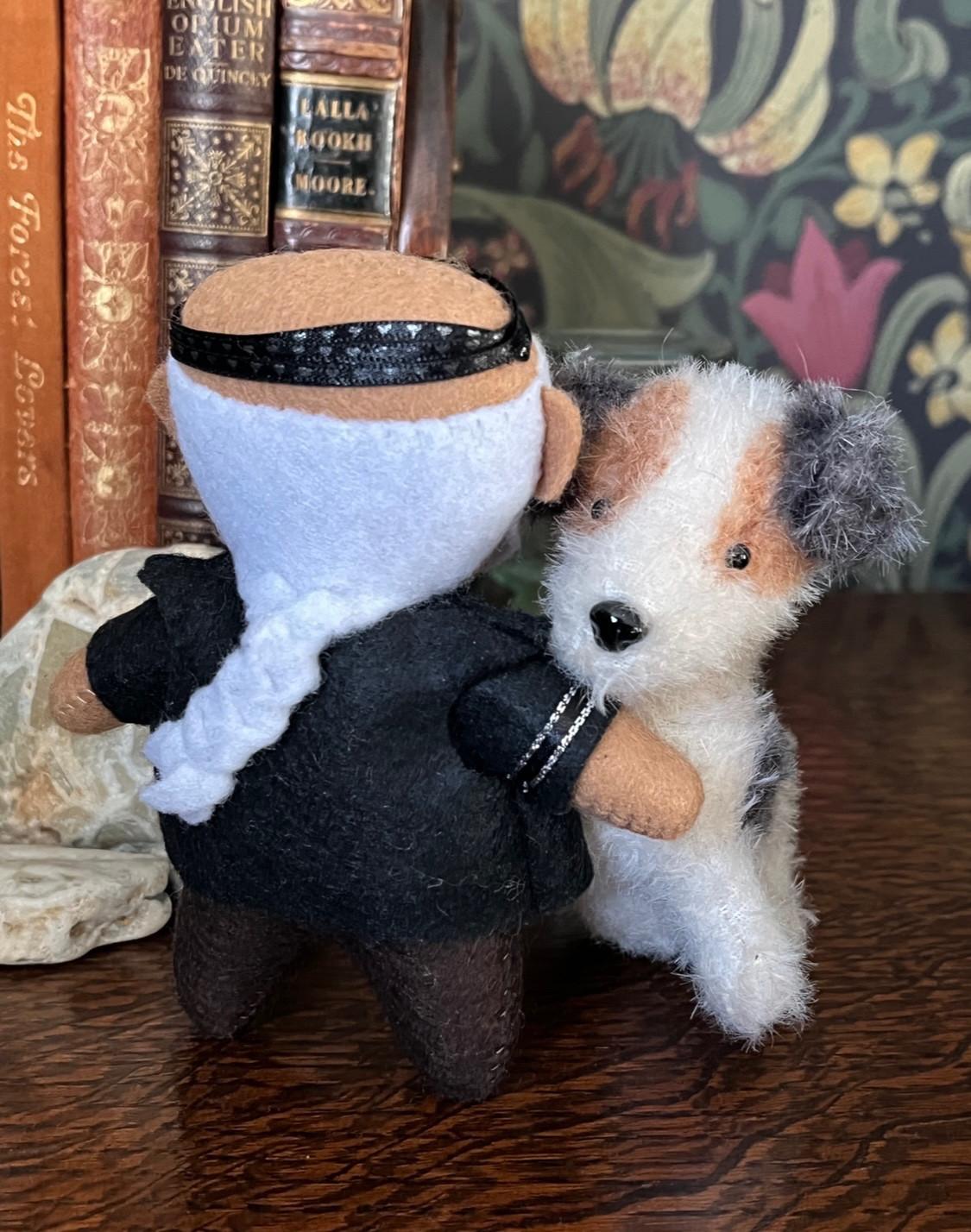 Small felt plush doll of Fang from Our Flag Means Death with a plush dog that looks like a Jack Russell - white with a brown eye patch and black ears. This is a back view of Fang hugging the dog so you can see his long white braid and studded head-band.