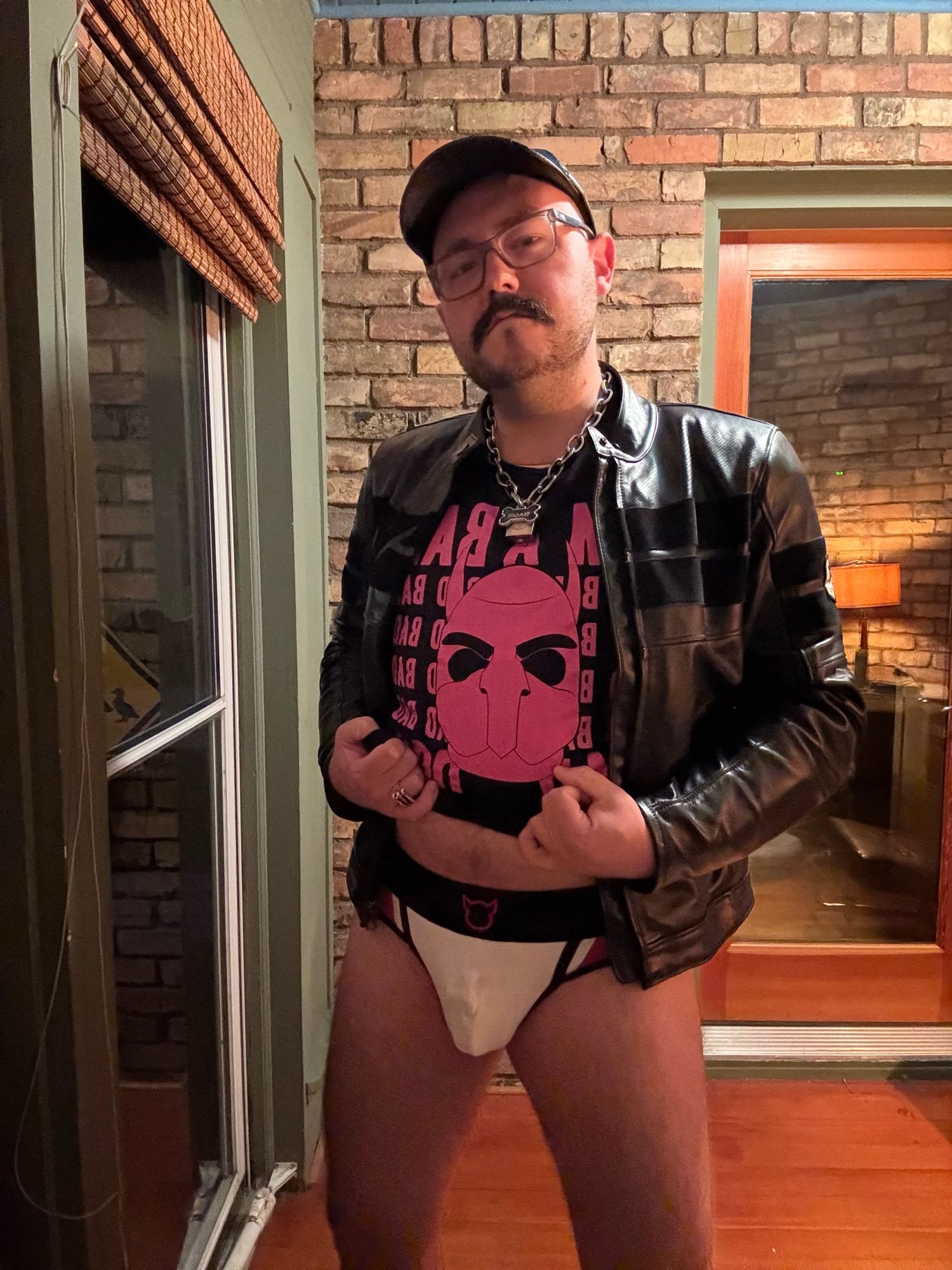 Faust wearing a leather motorcycle jacket with his pants down to reveal a white jockstrap with black and red detailing. His chastity cage imprint is visible through the jock.