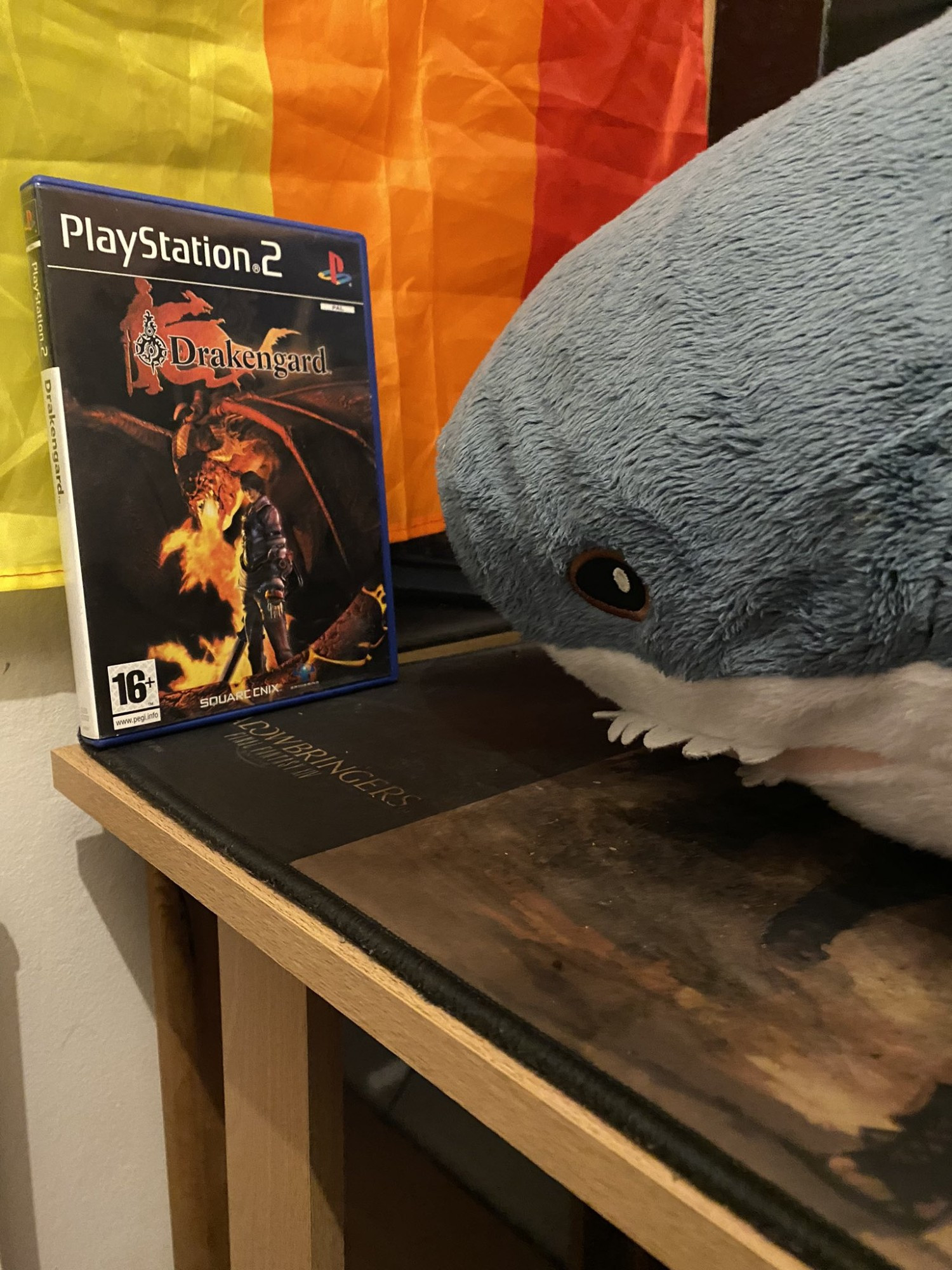 Picture of Blåhaj looking at a copy of Drakengard, with a Pride flag in the background