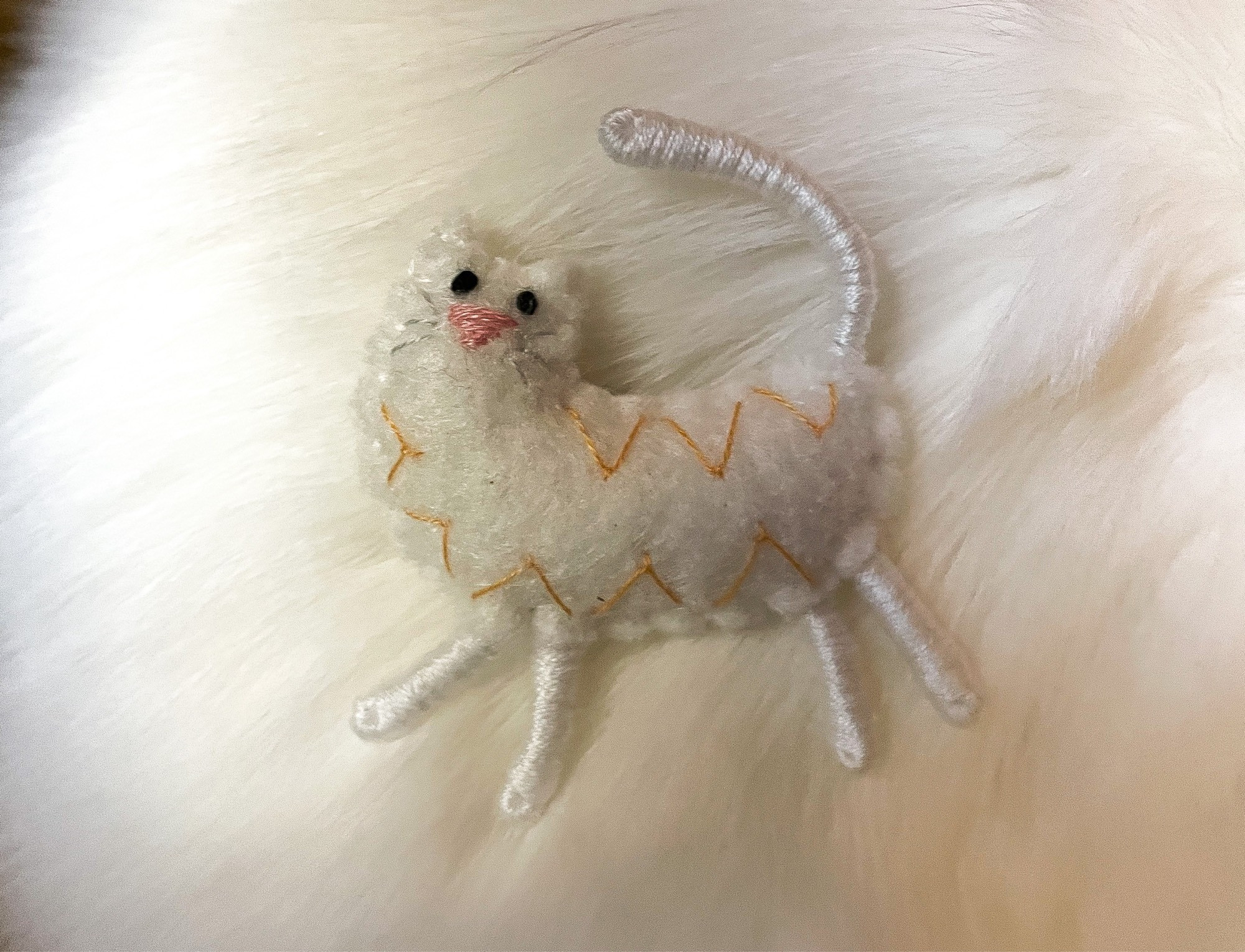 Photo of a small cartoonish white with a pink nose made from a lightly stuffed fabric body and embroidery thread wrapped wire legs. It is lying on a backdrop of my white cat Francie’s fur.