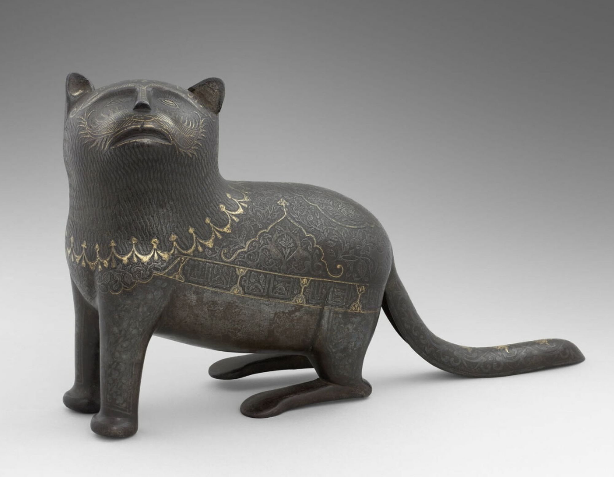 Very nice modern studio photograph of a metal cat sculpture. It is stylized and almost cartoonish but in an artful way. It is made from steel that is blackened and decorated with delicate gold ornamentation. The steel seems matte at first but closer examination reveals it is beautifully detailed with fur texture and floral designs. The cat it in a half- standing sort of crouch, looking upward with a somewhat humanoid face, as if begging for food from above.