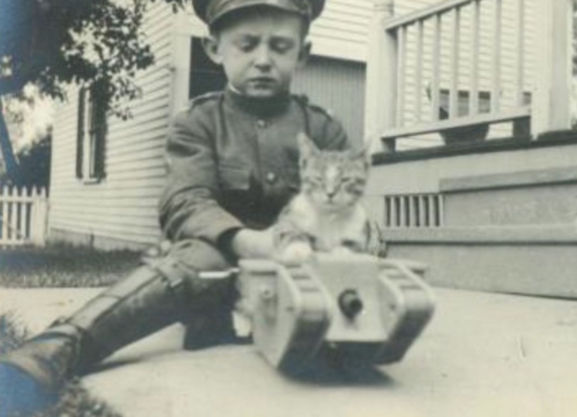 Black and white photo of a very serious looking little white boy in a child-sized WWI uniform and high leather boots. He's sitting on a sidewalk in front of a porch pushing a little white and tabby kitten on a toy tank. The kitten seems relaxed but disinterested.