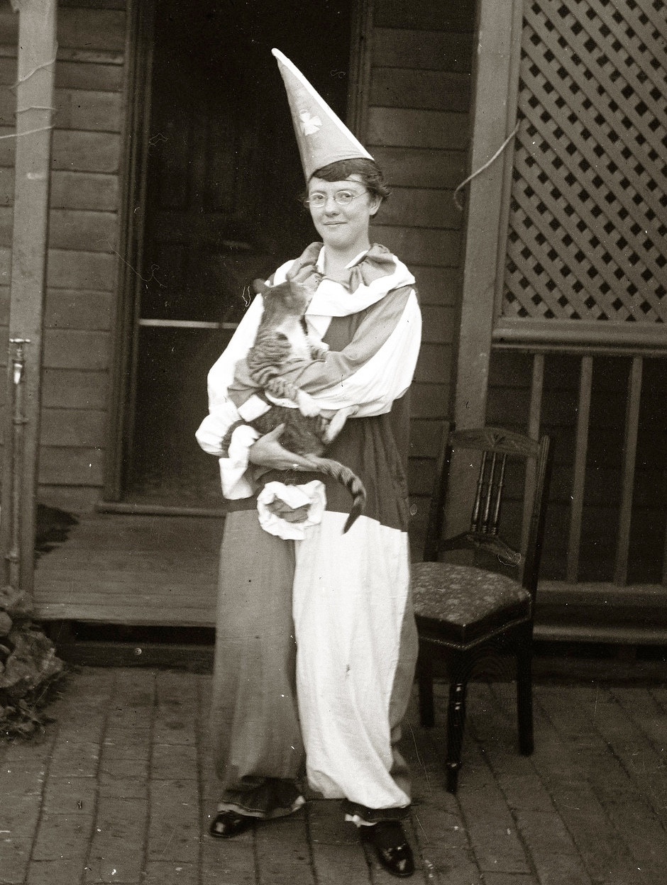 Black and white photo of a woman wearing a voluminous, baggy clown costume that is a jumpsuit made of large blocks of different colored fabric, a large, ruffled collar, and a cone-shaped tall hat. She is standing on a wooden porch and smiling at the camera. In her arms she cradles a shorthaired tabby cat whose head seems to be turned toward her face.