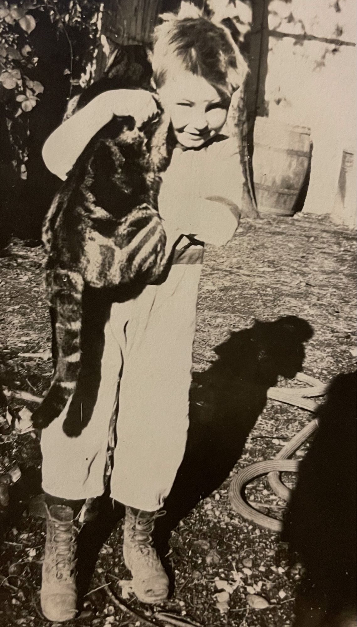 Black and white photo of a young white boy standing in a yard in the sun, smiling at the camera and awkwardly holding a large shorthaired tabby cat.